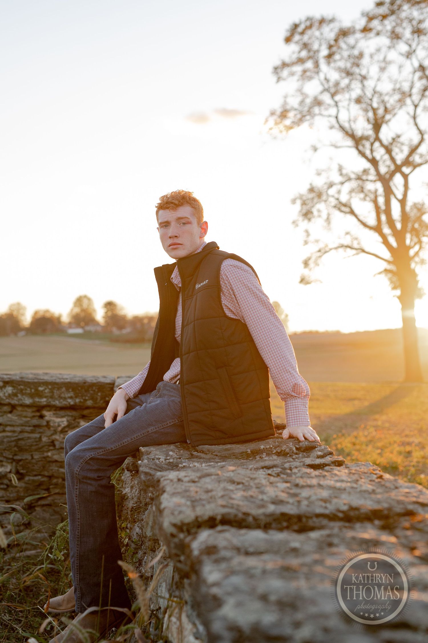 Person in dark vest sits casually on stone wall during sunset in rural setting.