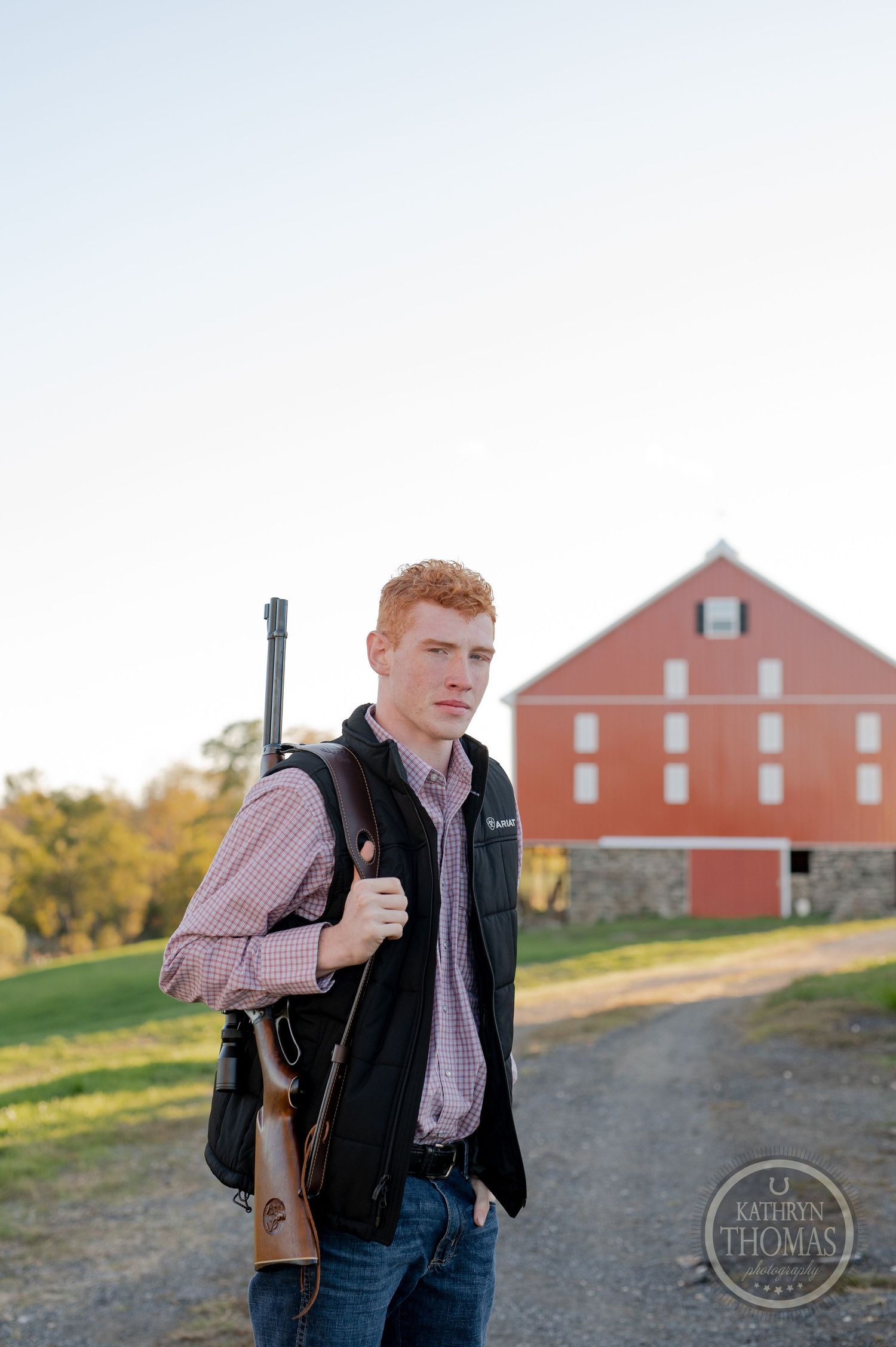 Person carrying rifle with red barn in background during golden hour countryside photoshoot.