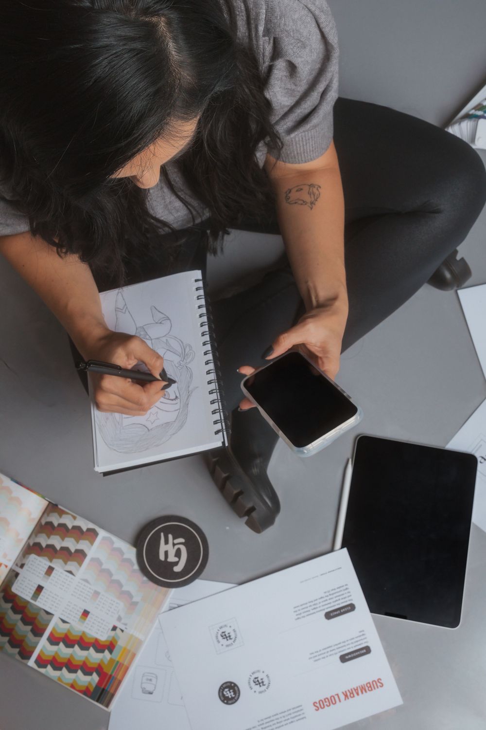 Overhead view of person sketching logos while working with mobile device and business materials.