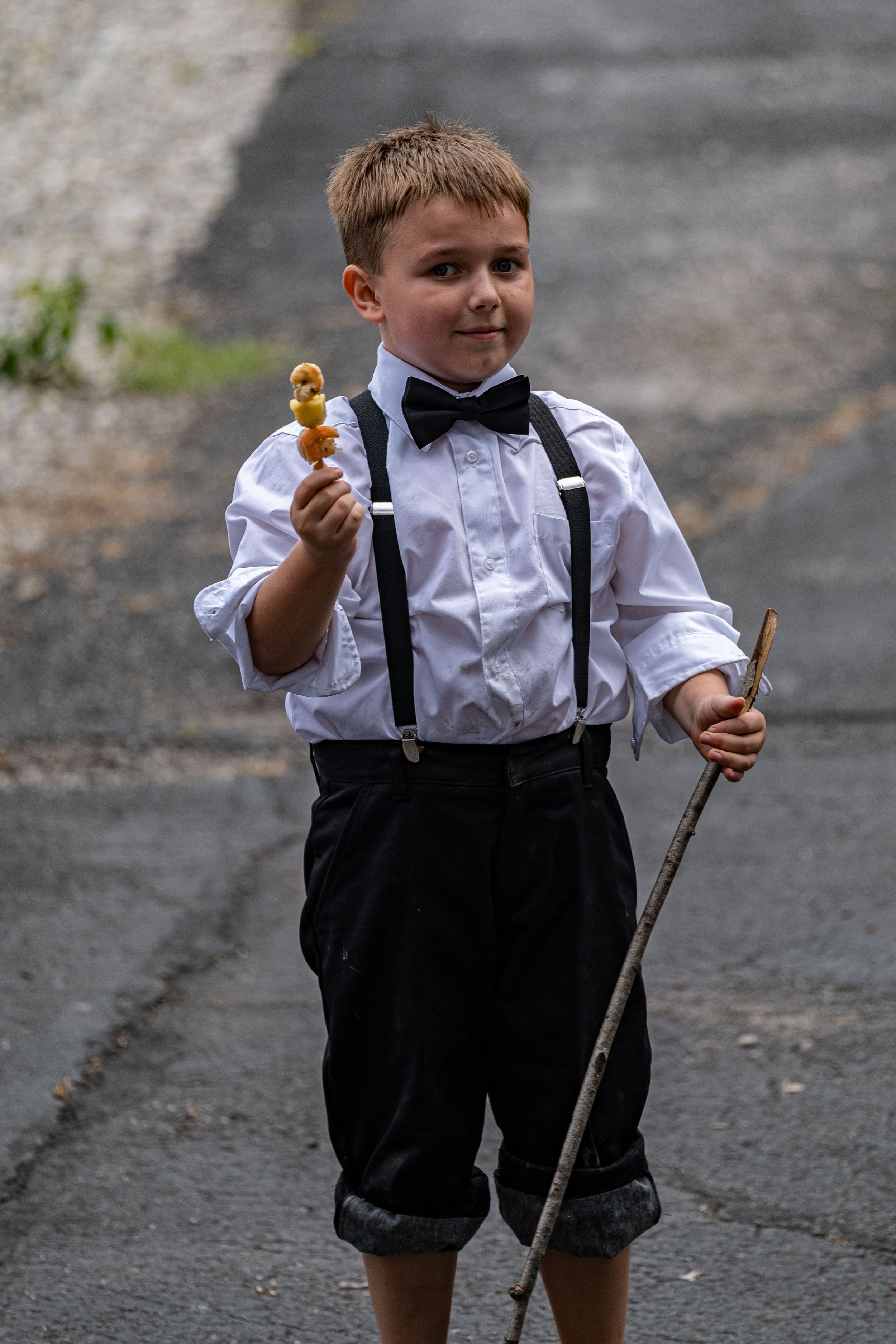 Kid in tuxedo and suspenders poses for picture with his walking stick and kabob.