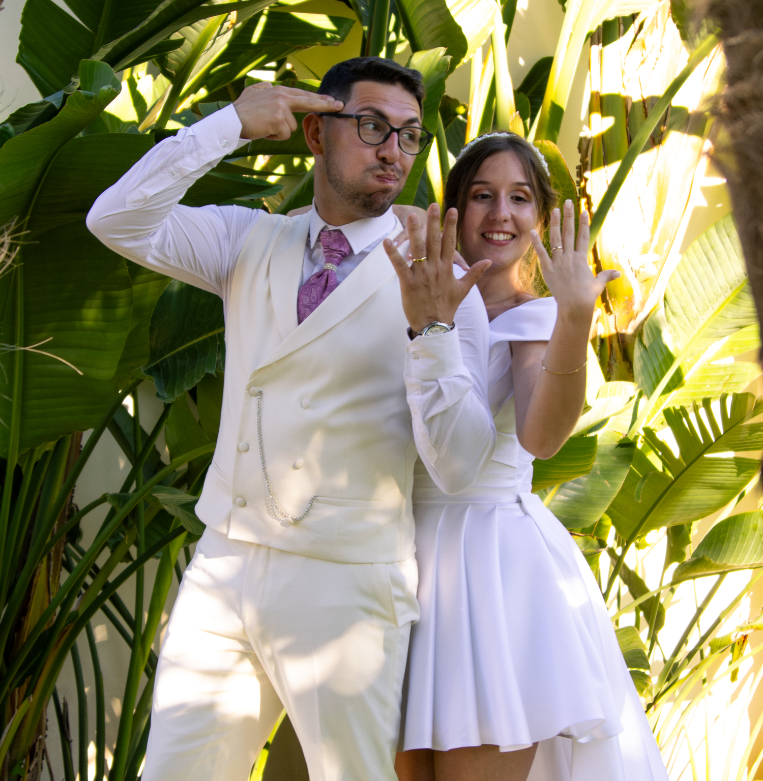 A couple in white attire poses playfully showing off their rings against tropical foliage.