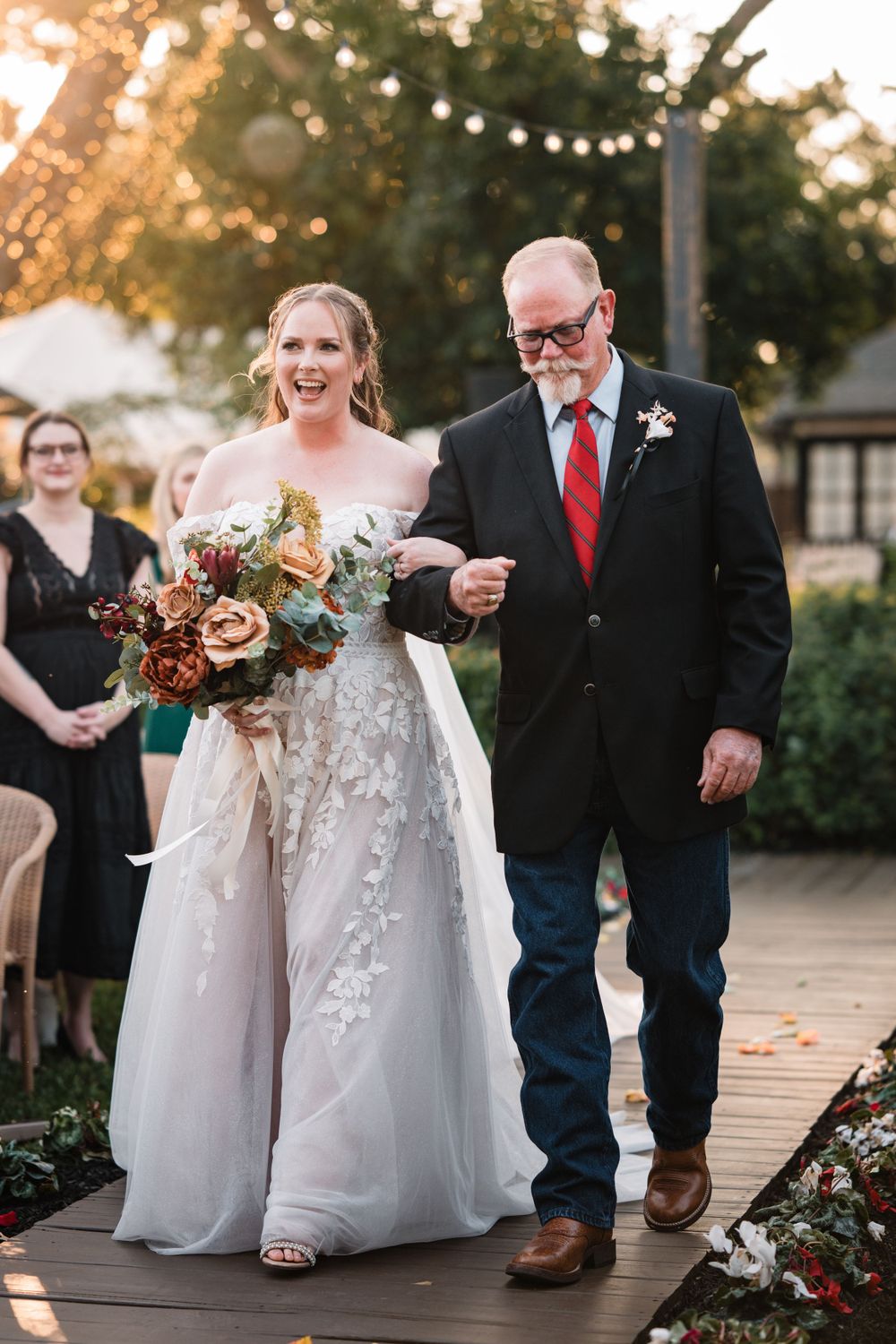 Wedding party walking down outdoor path with bride in white dress and autumn floral bouquets.