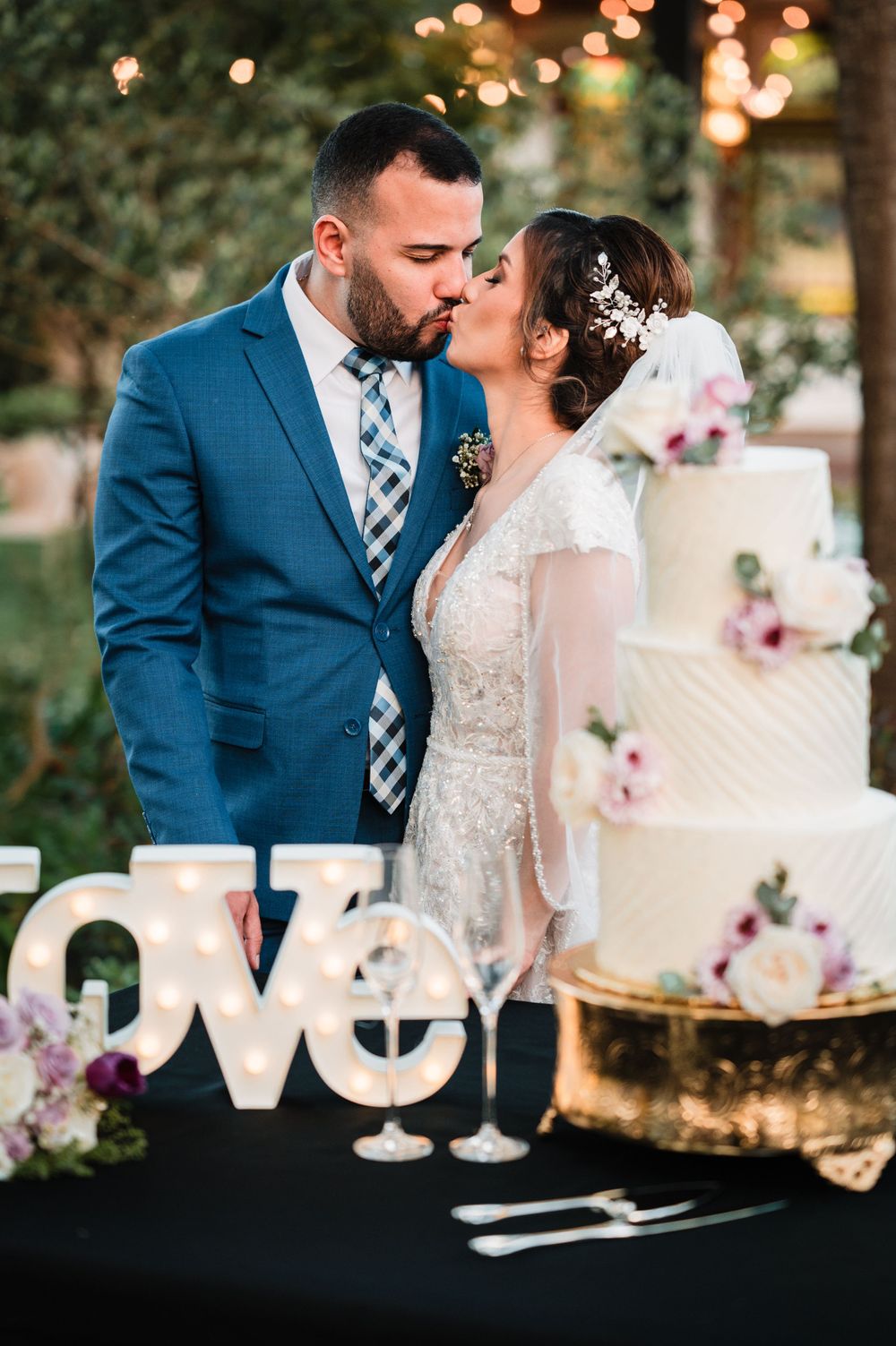 Elegant white wedding cake display with floral accents and 'love' sign at sunset reception.
