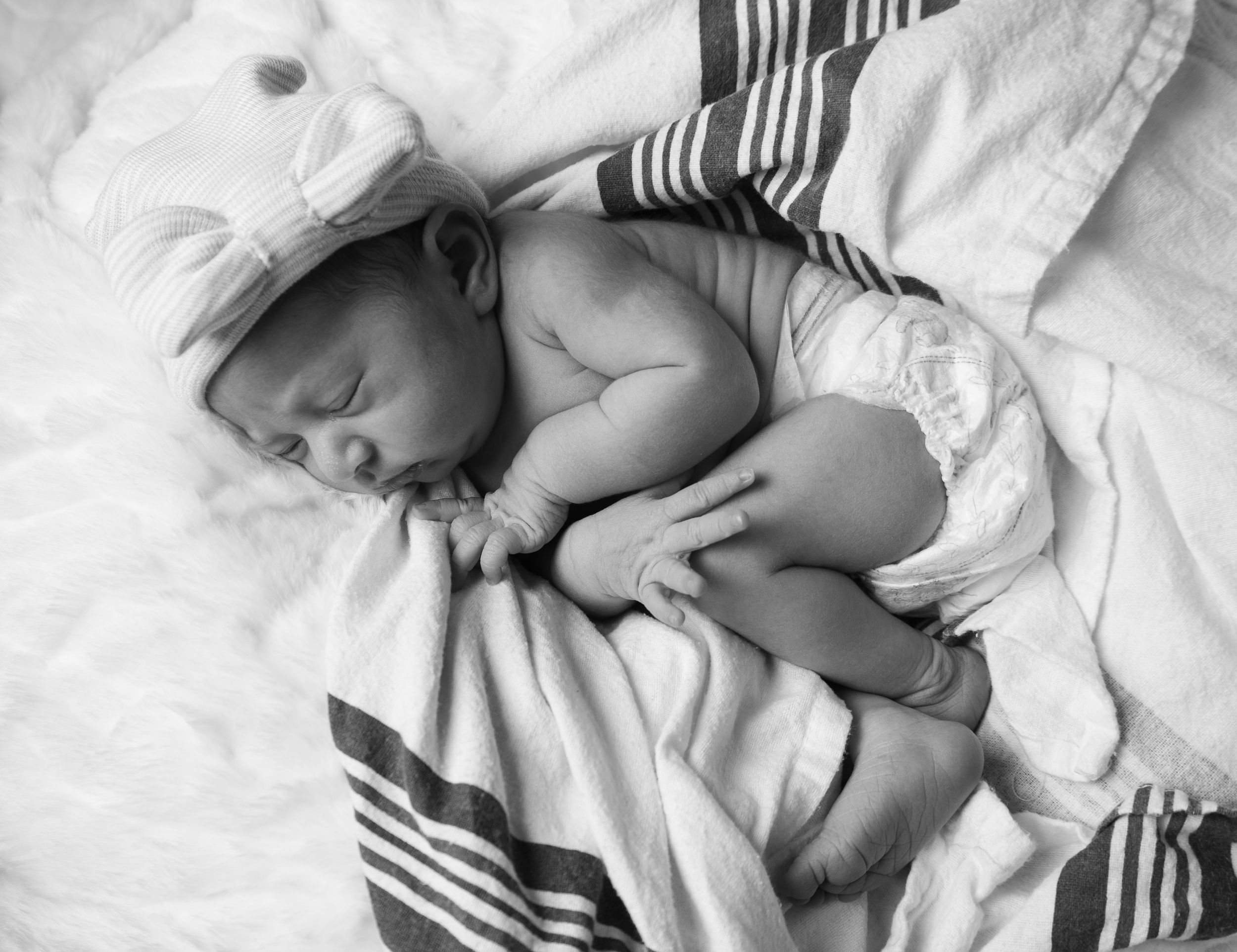 Peaceful newborn sleeping on white blanket with striped fabric and small white cap.