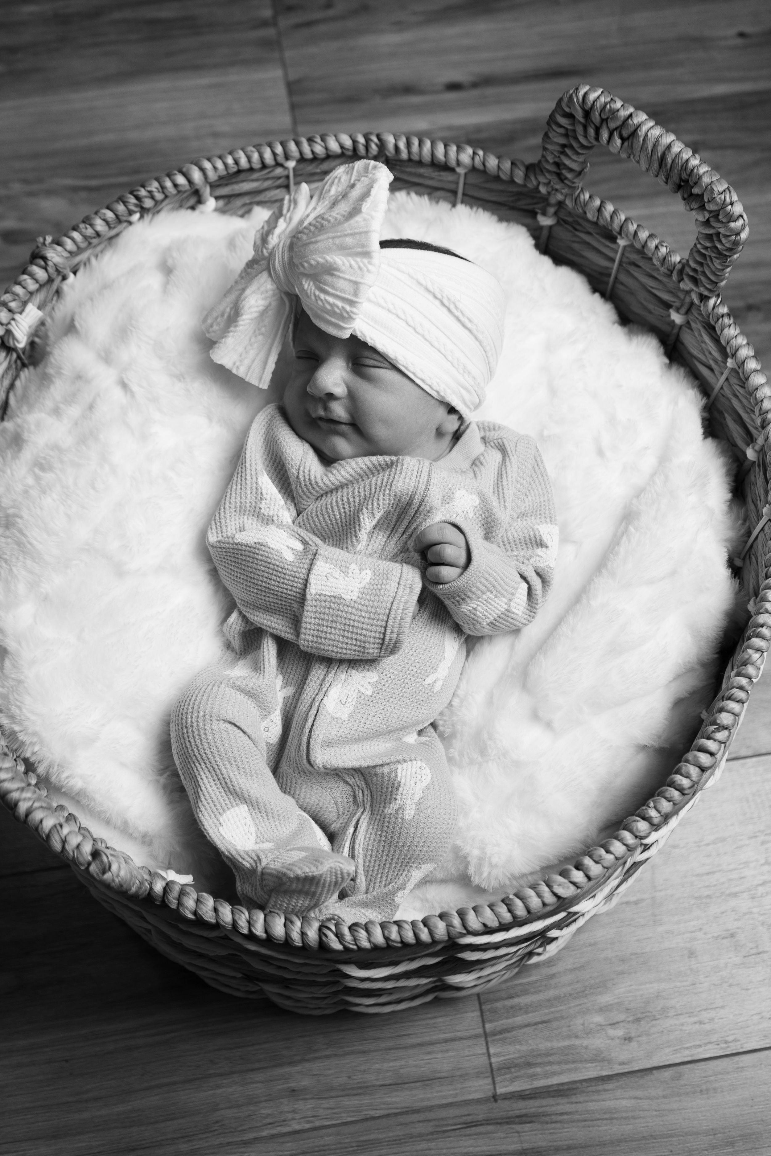 Sleeping newborn in white bow headband and pink outfit resting in wicker basket on white blanket.