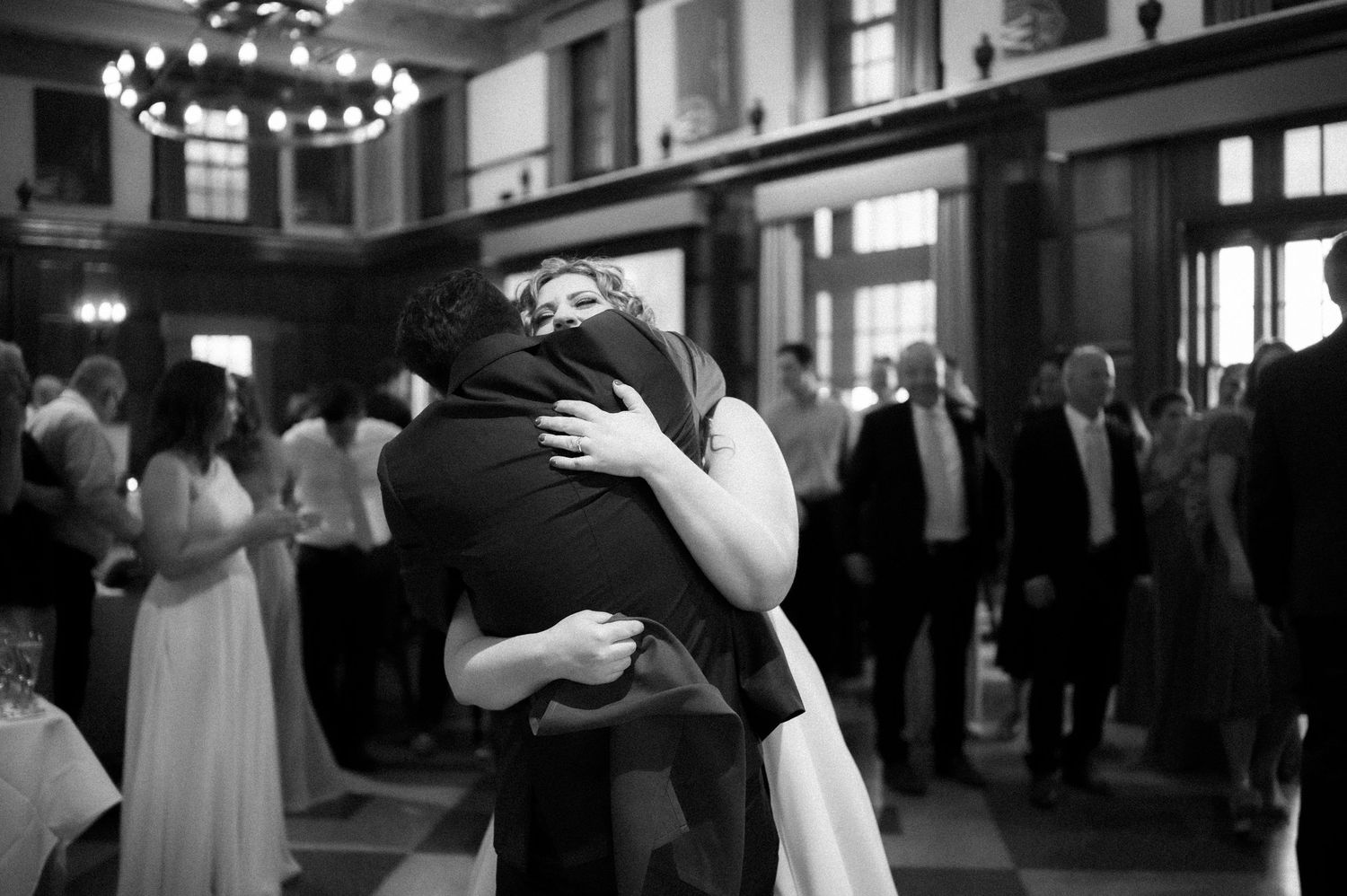 Two people share an intimate embrace on a dance floor while others gather in a dimly lit ballroom.