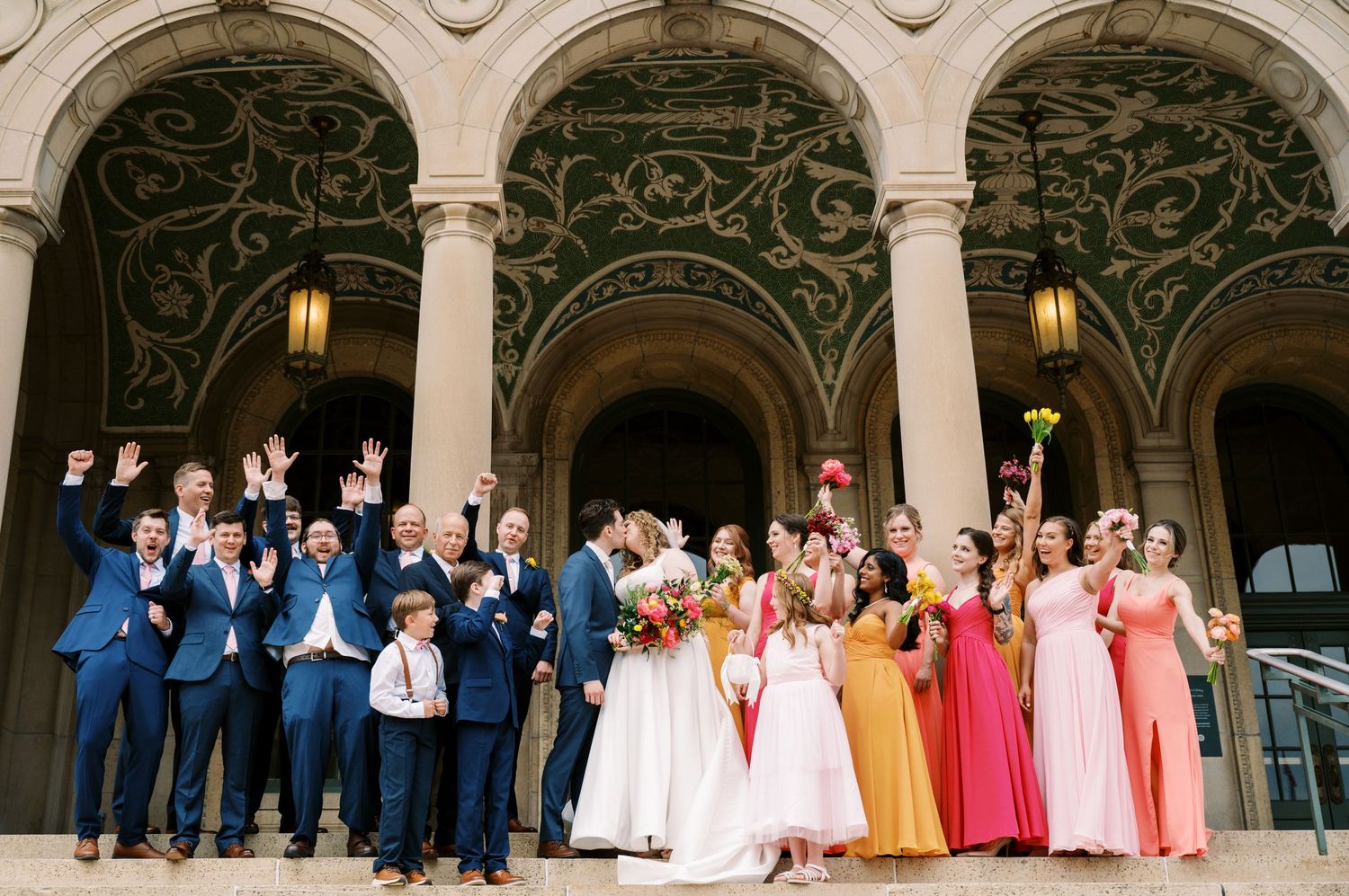 A wedding party poses under ornate stone arches with bridesmaids in colorful dresses and groomsmen in navy suits.