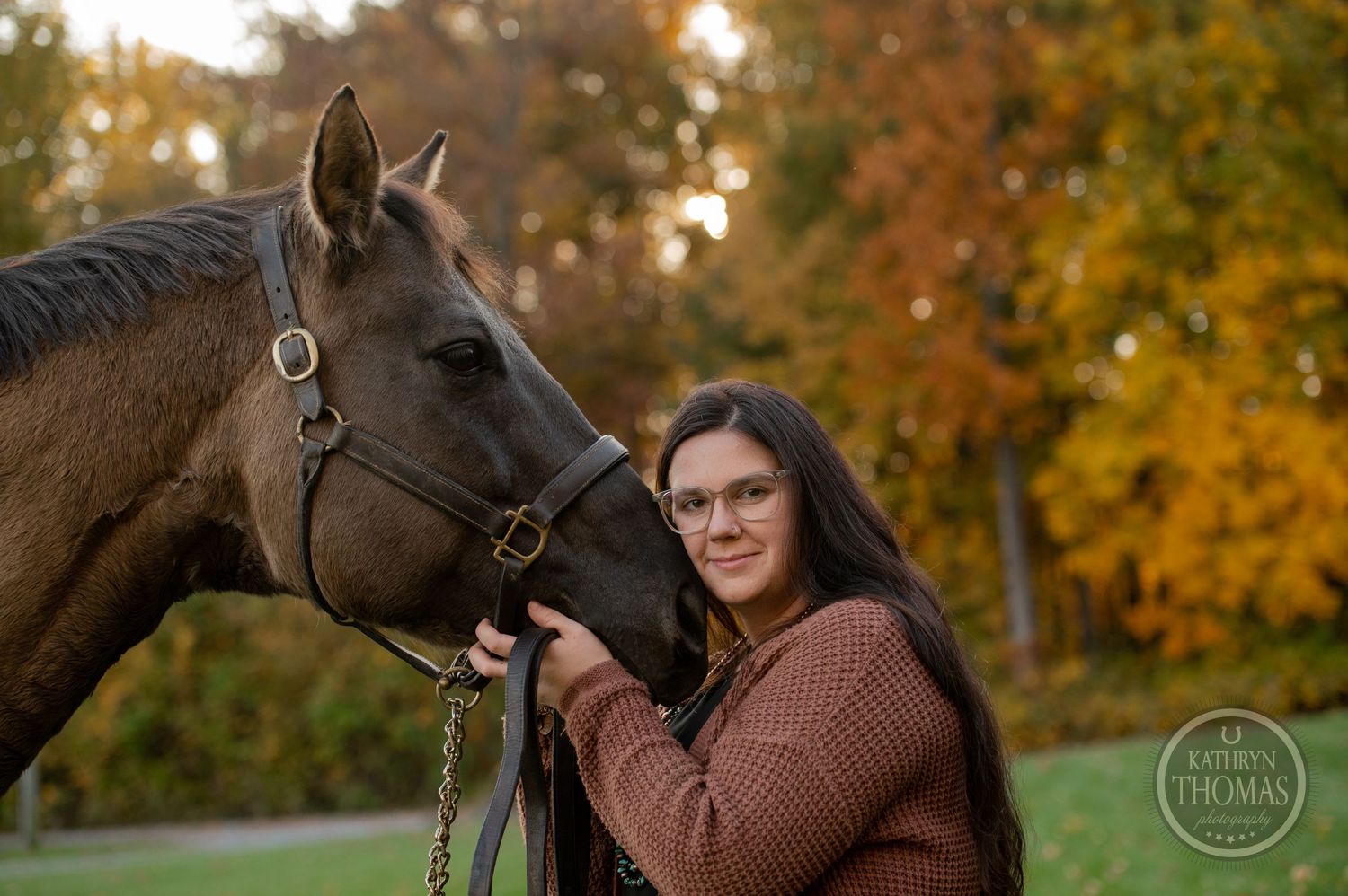 A brown horse and person pose together on a hill with autumn trees creating a warm backdrop.