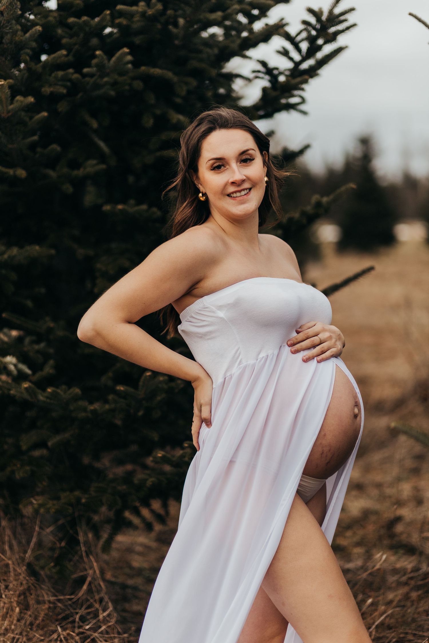 A maternity photo taken outdoors featuring a flowing white dress amidst evergreen trees at sunset.