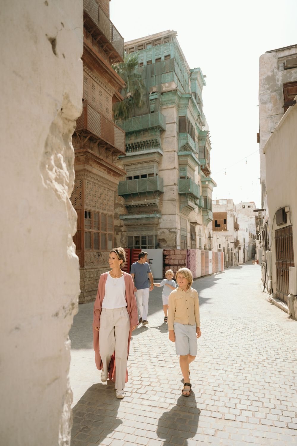 A woman and child walk through a historic cobblestone alley in Al-Balad, Jeddah, with ornate mashrabiya buildings in the background.