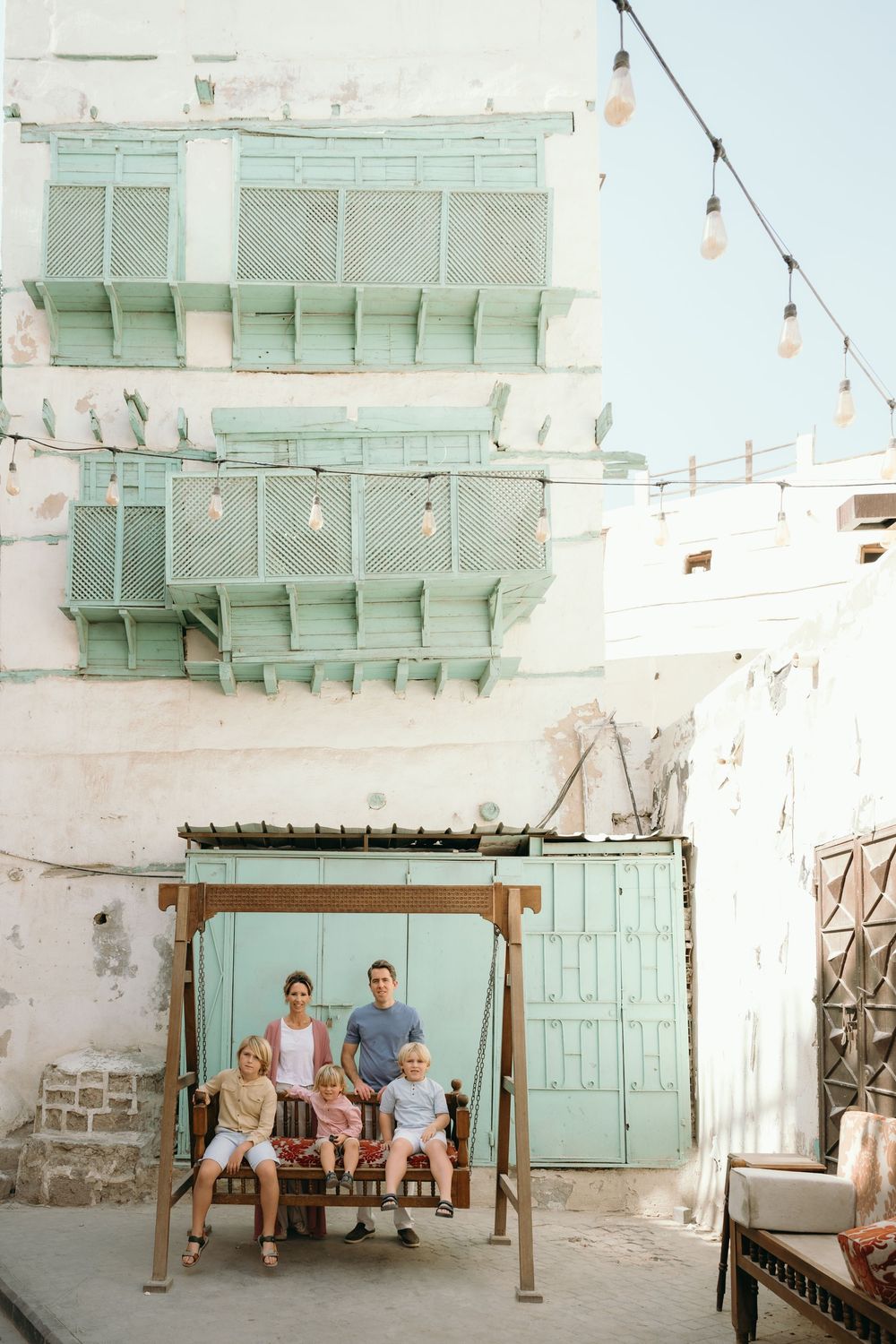 A family of four sits together on a wooden swing in front of a historic white building with turquoise mashrabiya windows in Jeddah.