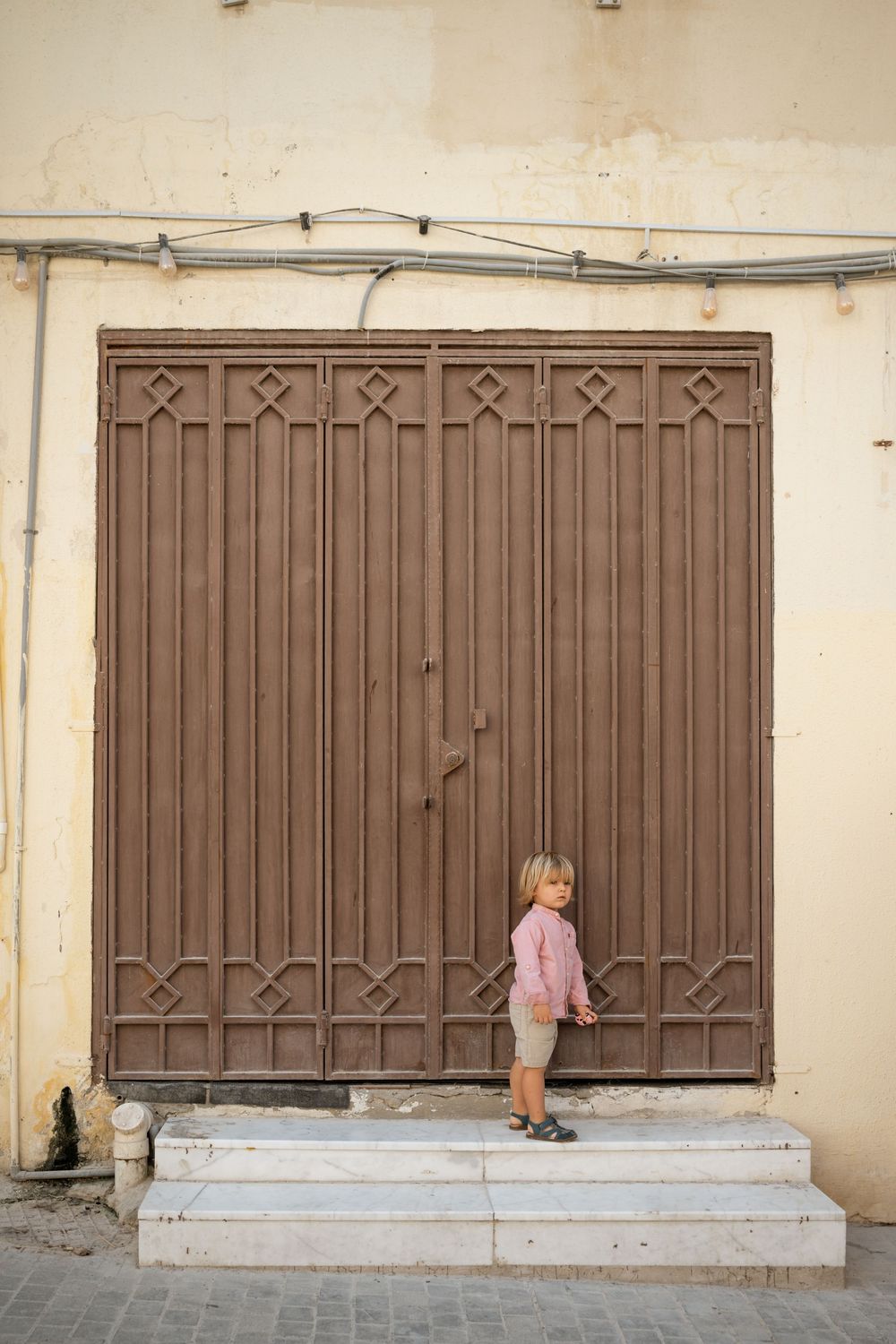 Young blonde toddler standing on white steps in front of large ornate brown metal doors on a beige wall.