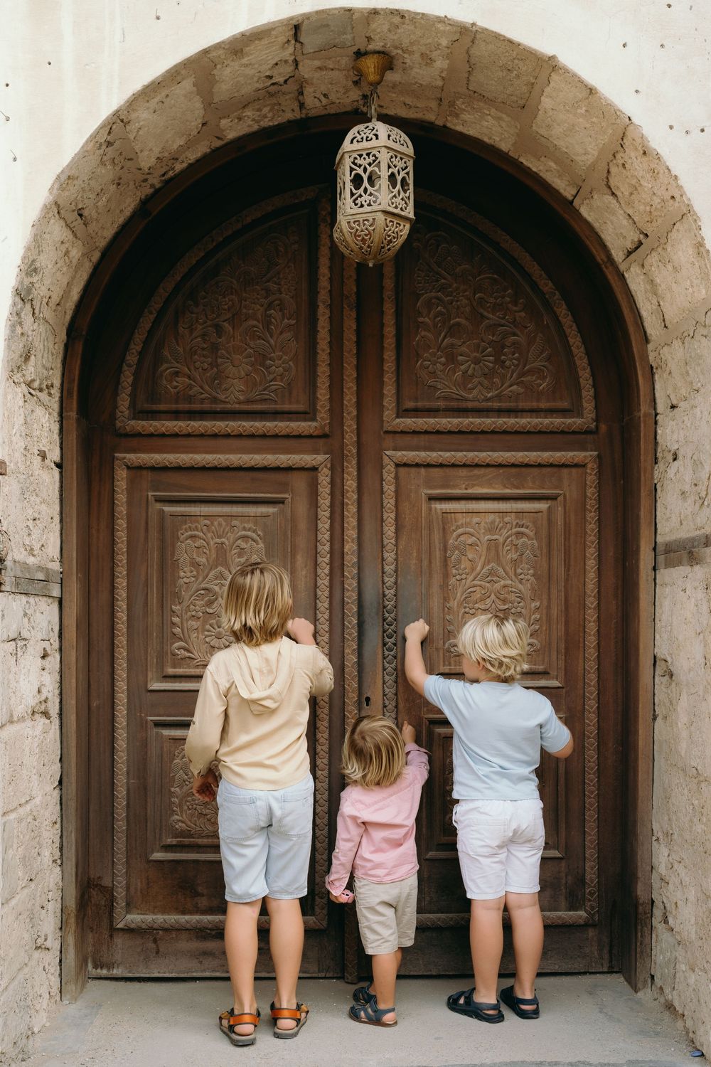Three children knocking on a large ornate wooden arched door with decorative carvings and a hanging lantern.
