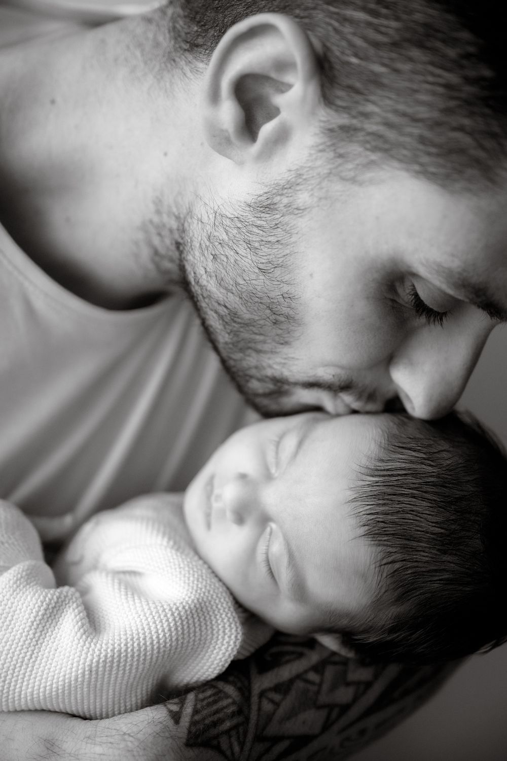 Black and white photo of a bearded father gently kissing his sleeping newborn baby wrapped in a soft blanket.