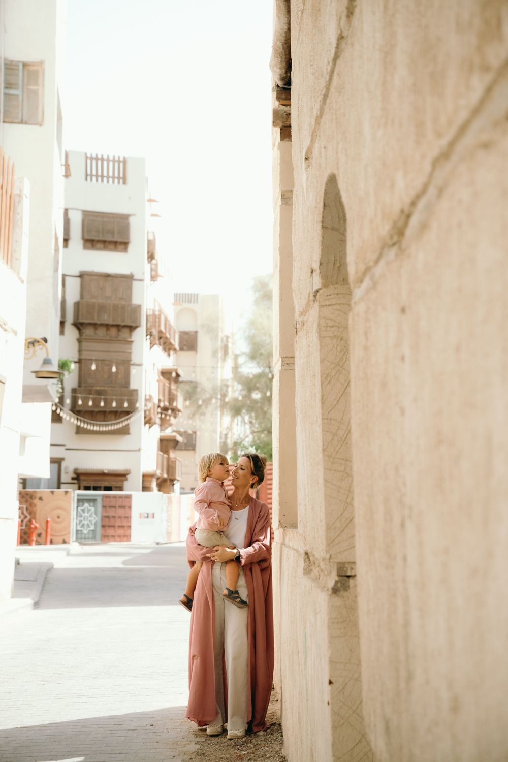 Mother in pink dress kissing young child while standing in a sunlit Middle Eastern alleyway.