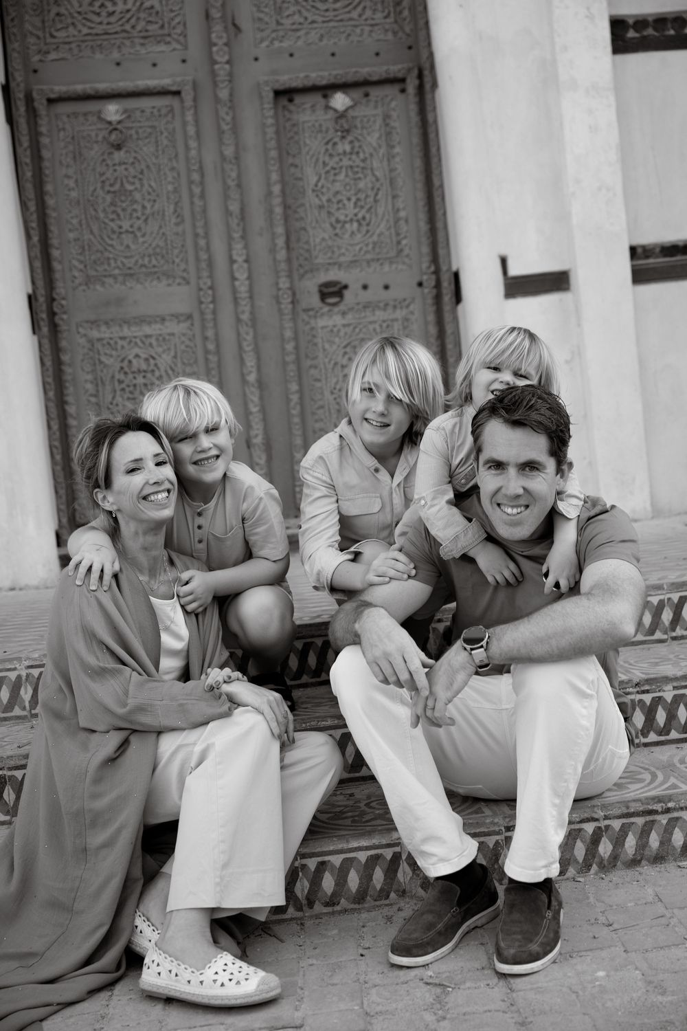 Black and white family portrait of parents and two children sitting on steps before ornate wooden door.