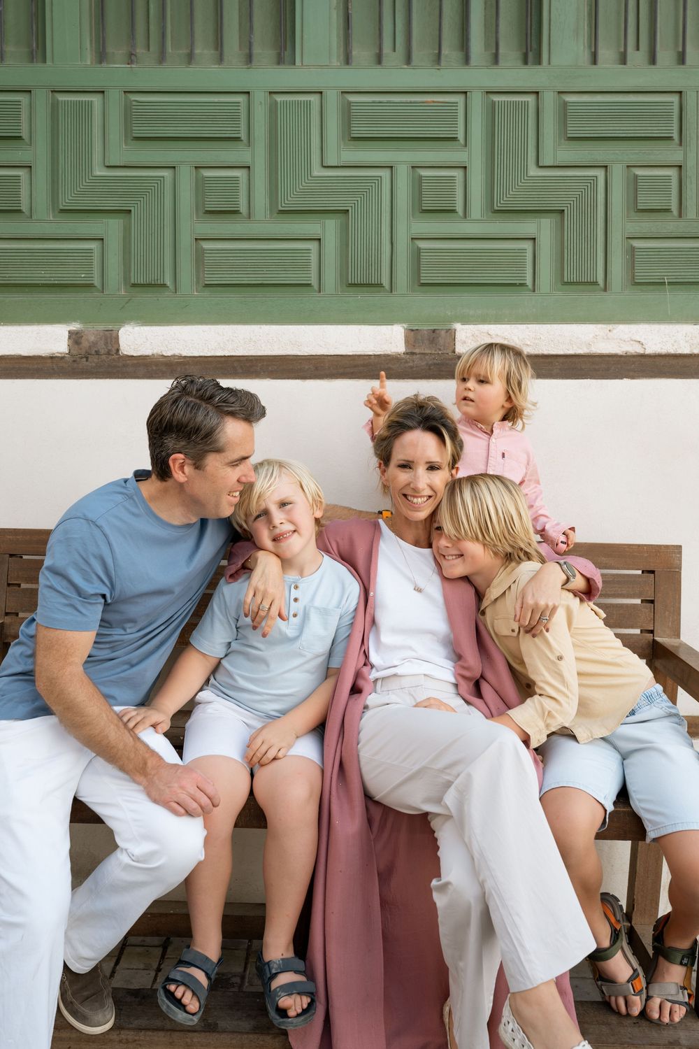 Happy family of five sitting together on a wooden bench, smiling and embracing outdoors near a decorative green wall.