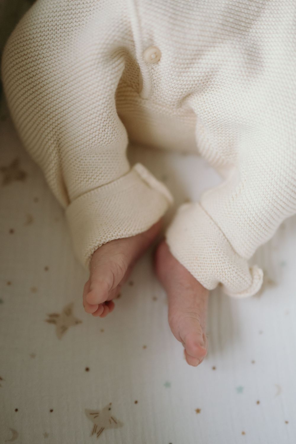 Tiny newborn baby feet peeking out from a cream knit onesie, lying on a white star-patterned surface.