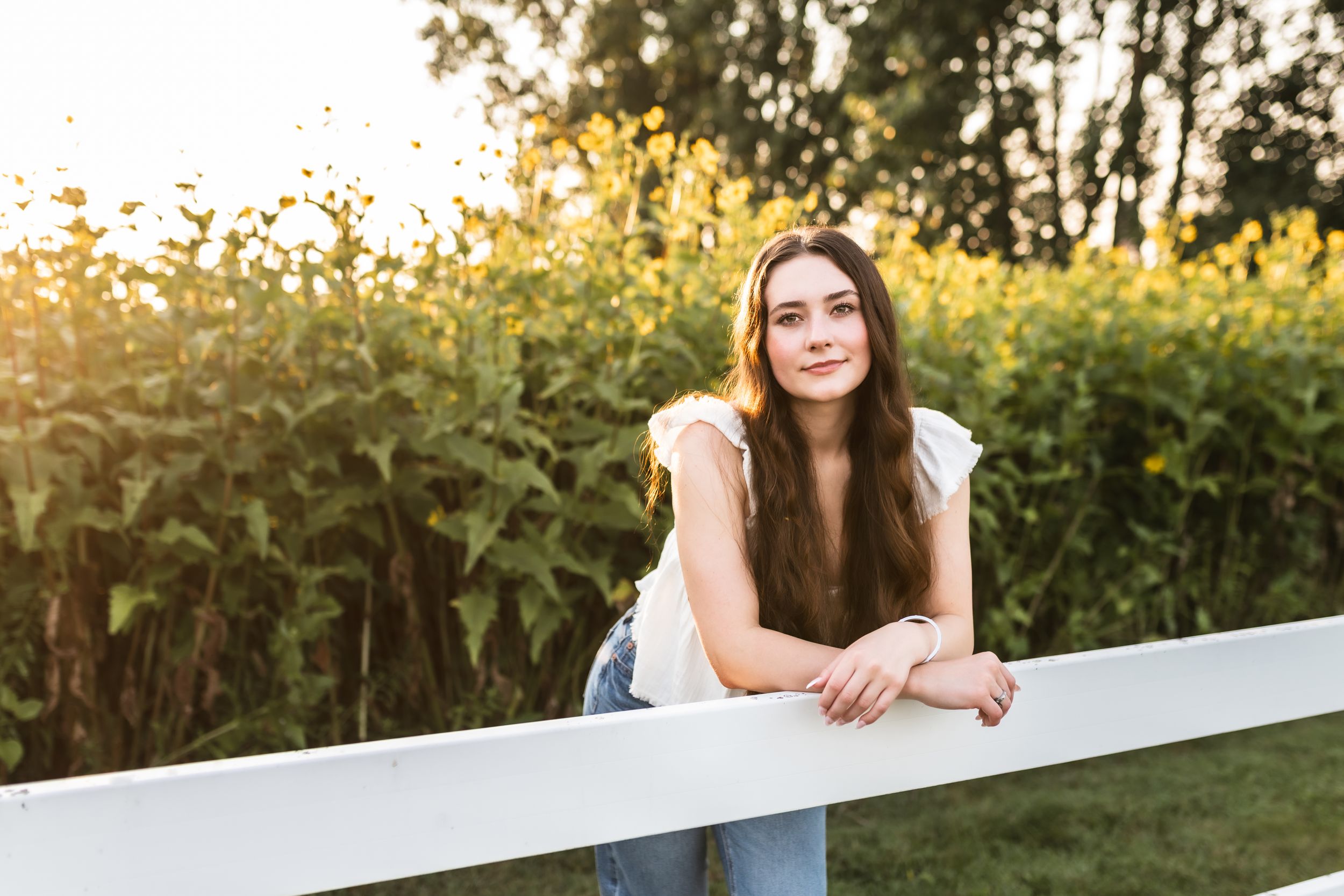 Golden Hour portrait of a senior girl leaning on a white fence with the sun glowing on the field behind her.