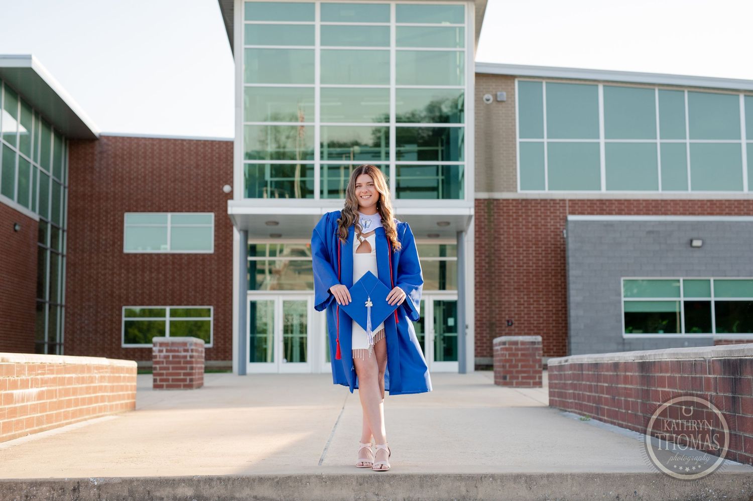 Graduate wearing blue commencement attire stands elegantly at school entrance.