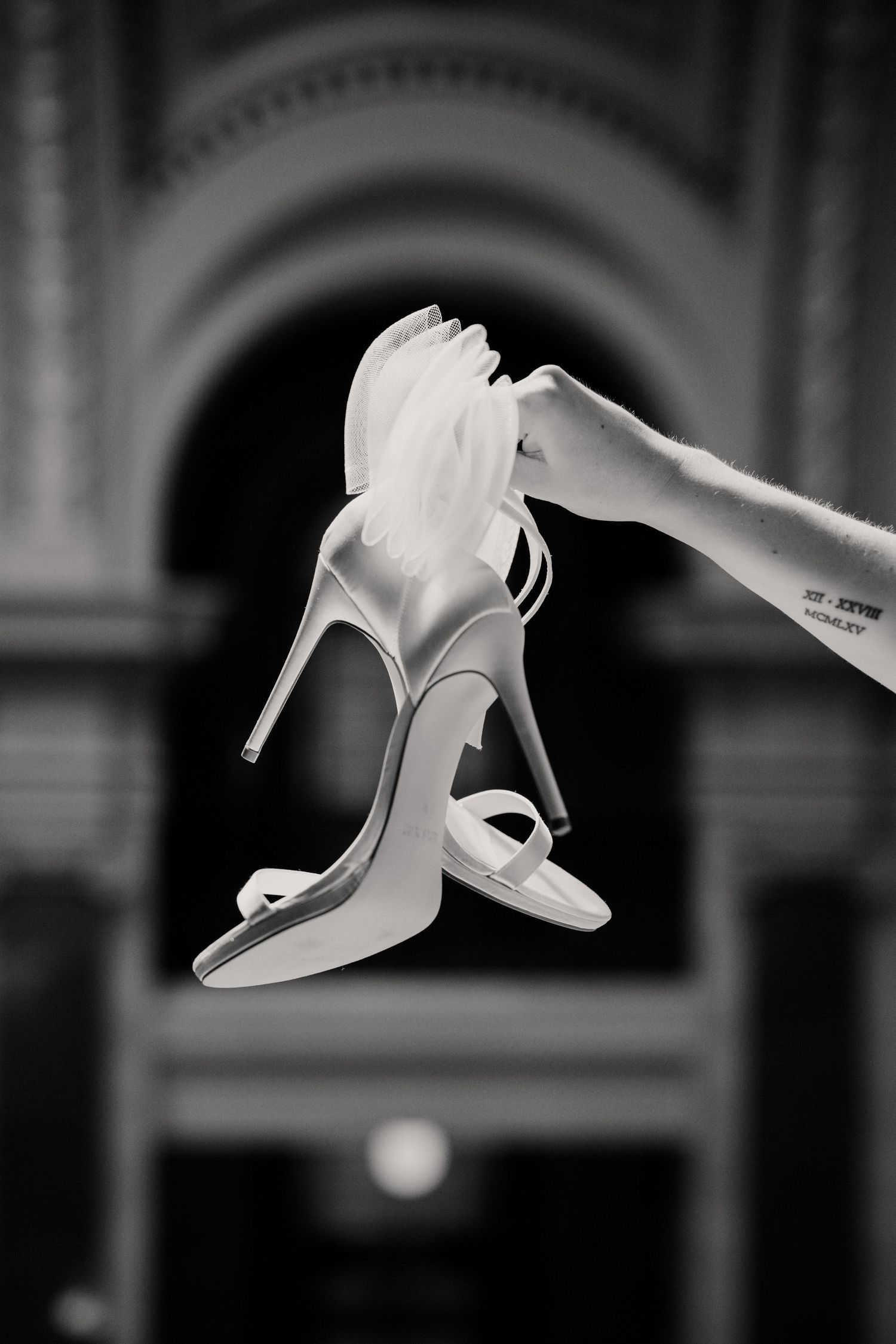 Hand holds white high heel bridal shoe in elegant black and white photo with architectural details in background.