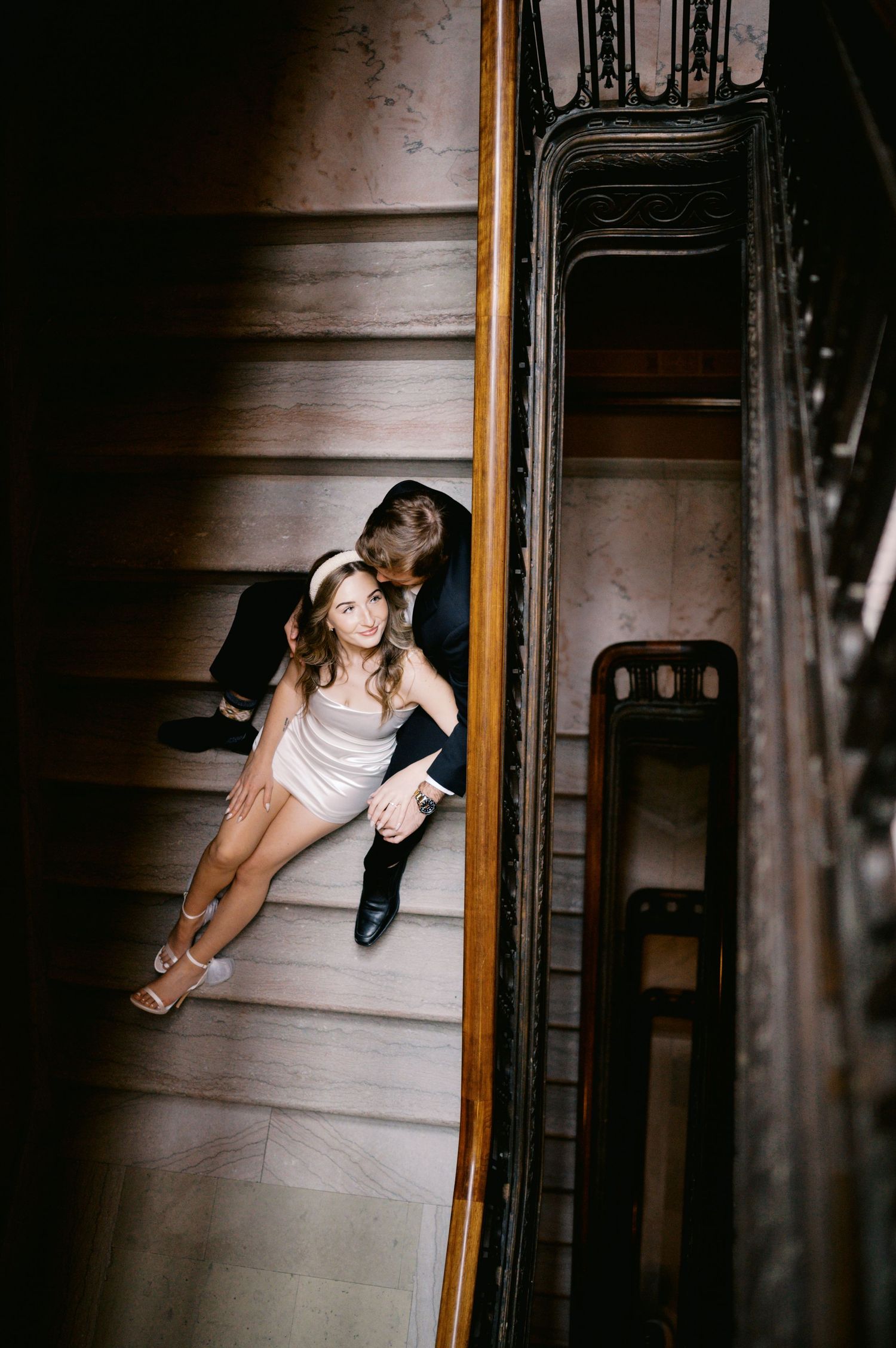 A couple sit together on wooden stairs in a vintage building with ornate banisters and dramatic lighting.