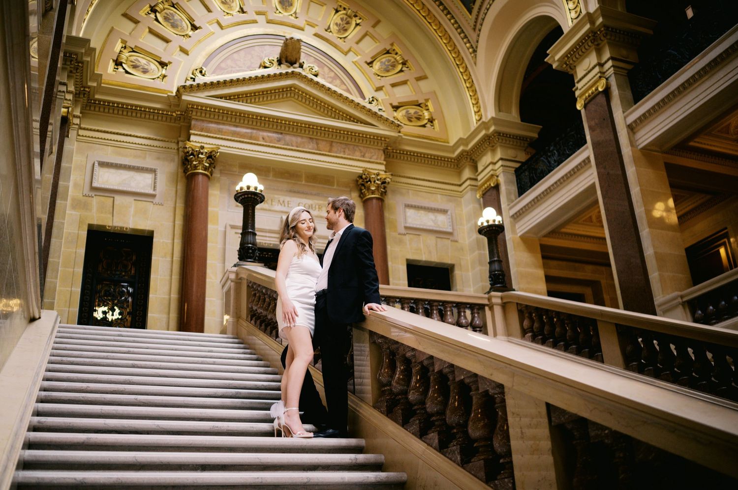 Couple stands together on grand marble staircase inside ornate historic building with elegant architectural details.