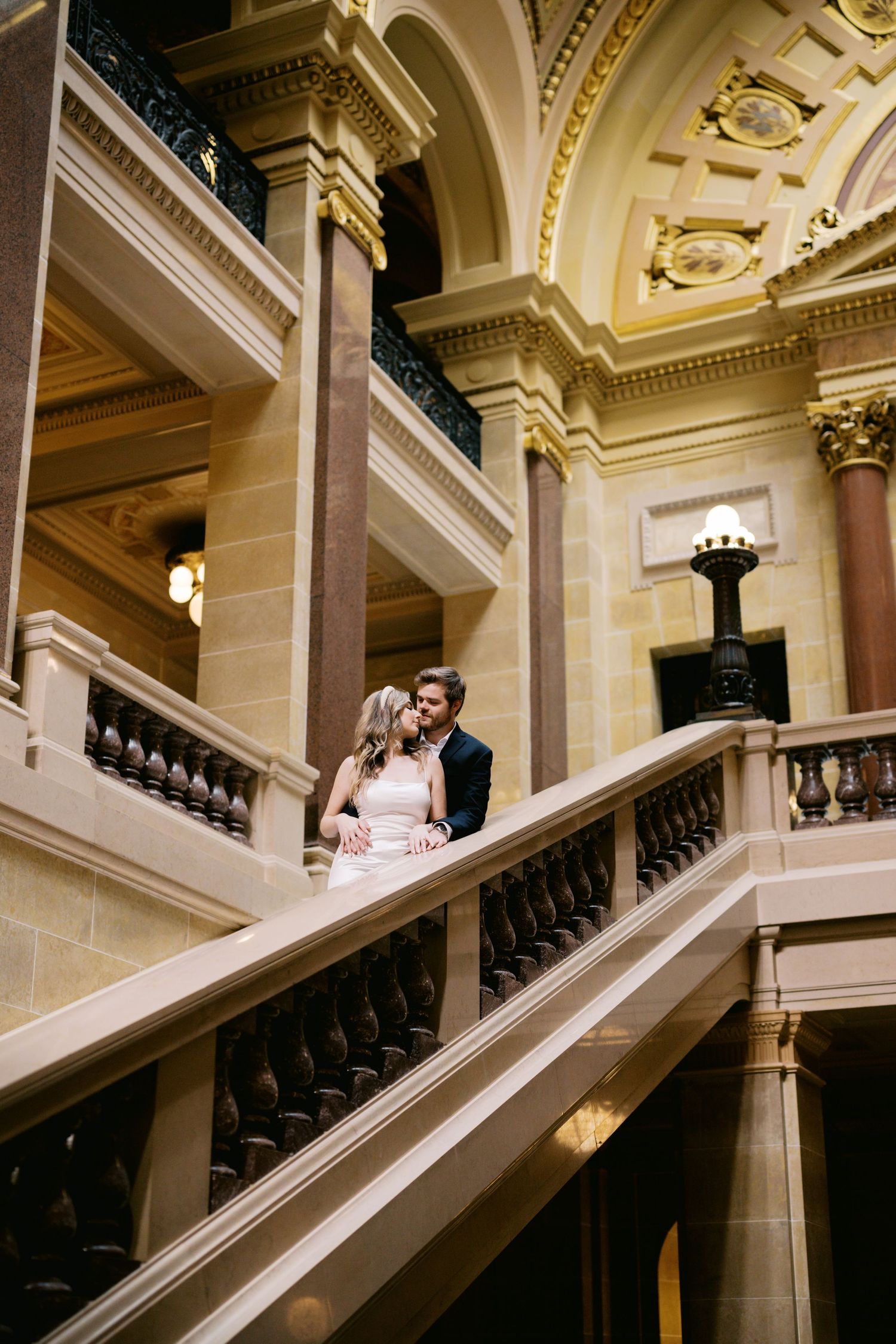 A couple stands together on an ornate marble staircase inside an elegant historic building with decorative architecture.