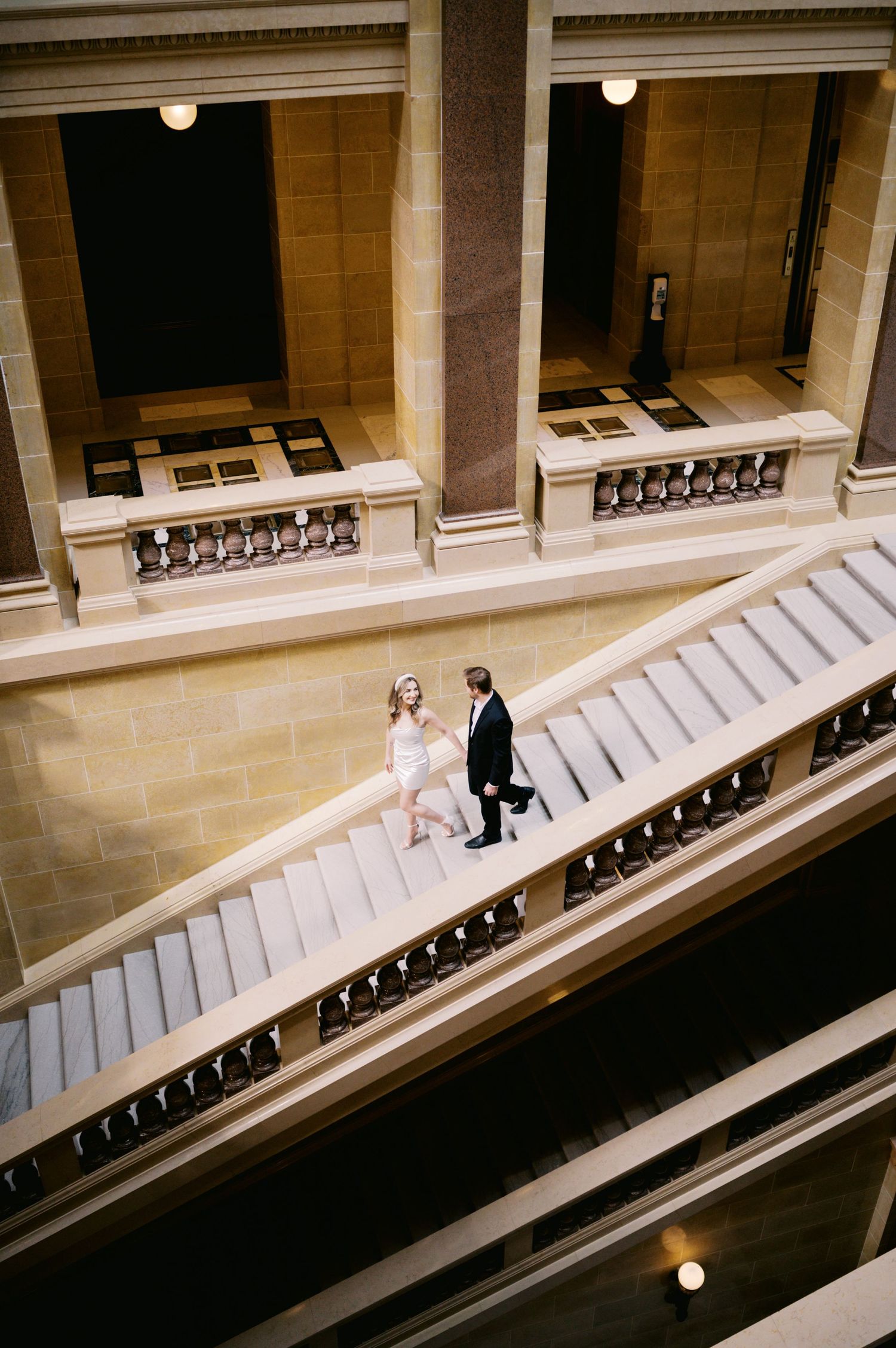 A couple in wedding attire walking down an elegant marble staircase with ornate columns and vintage architecture.