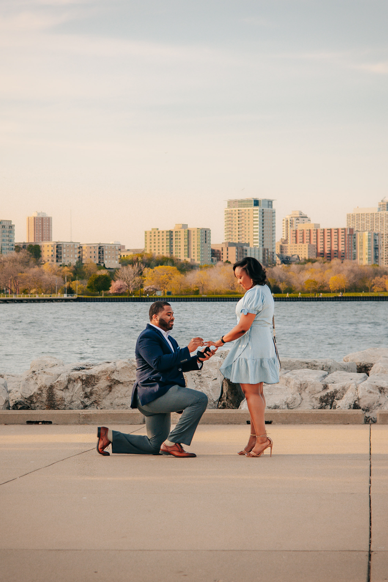Proposal moment at sunset by waterfront with city skyline in background.