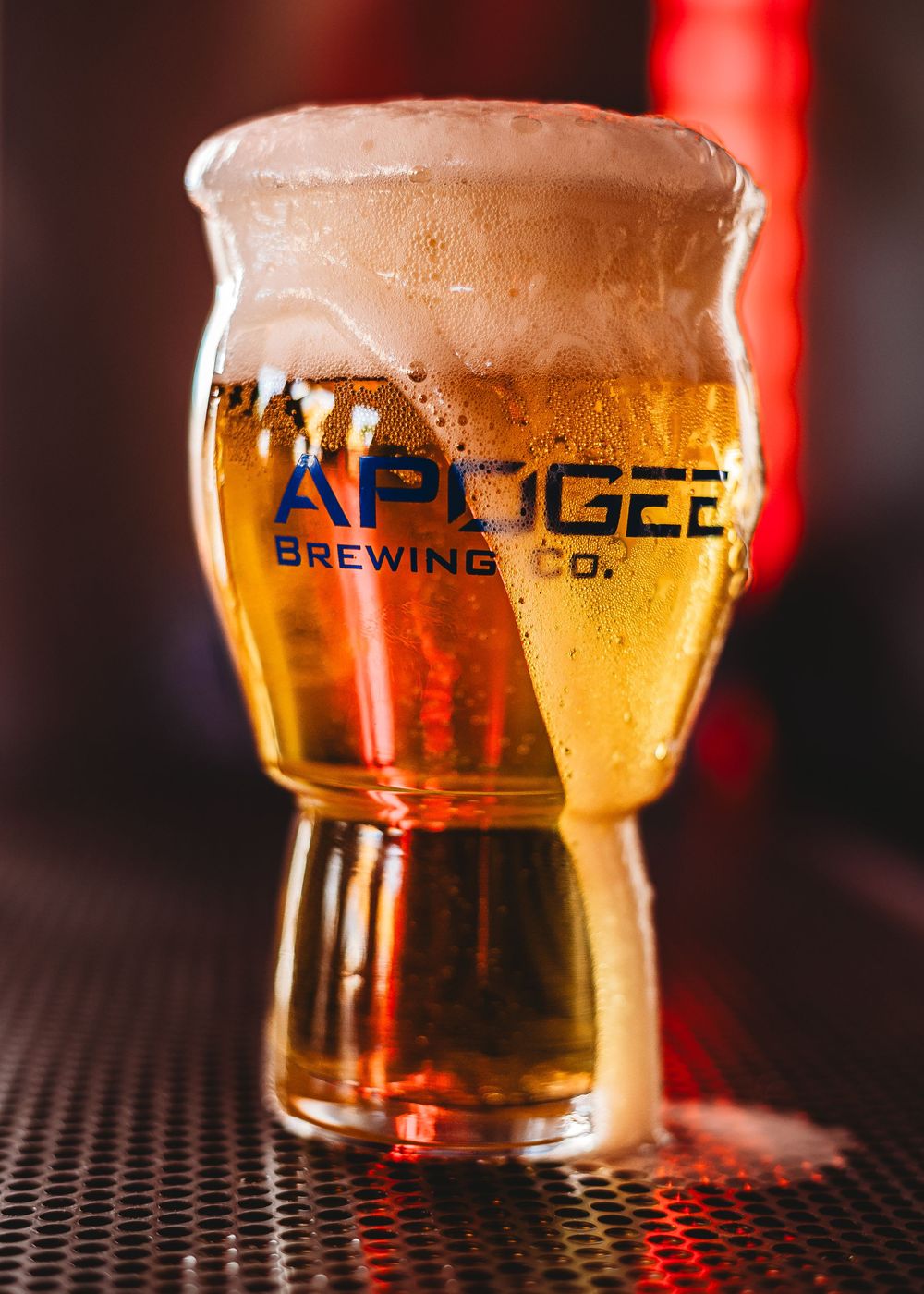 Apogee Brewing Co. logo etched pint glass filled with golden beer under red lighting and beer flowing out of the glass