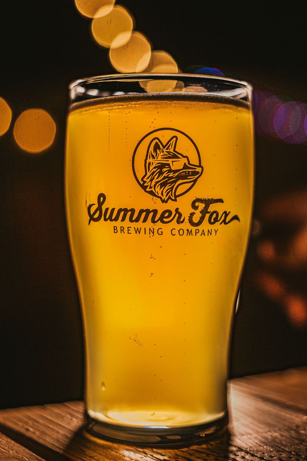 Beer glass with fox-logo pint glass filled with golden beer, string lights blurred in background