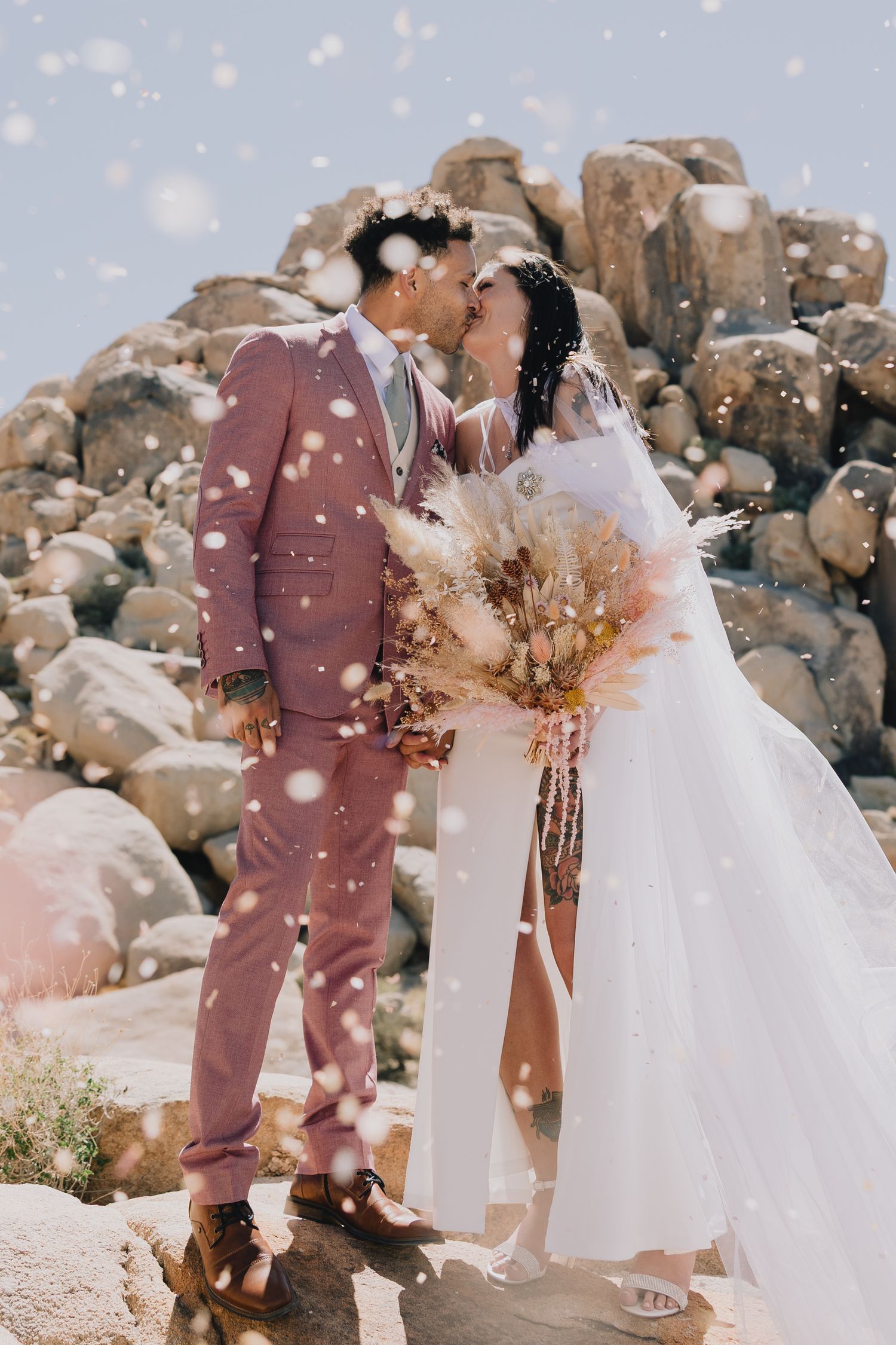 Bride and groom kiss surrounded by falling confetti against rocky desert landscape in Joshua Tree.