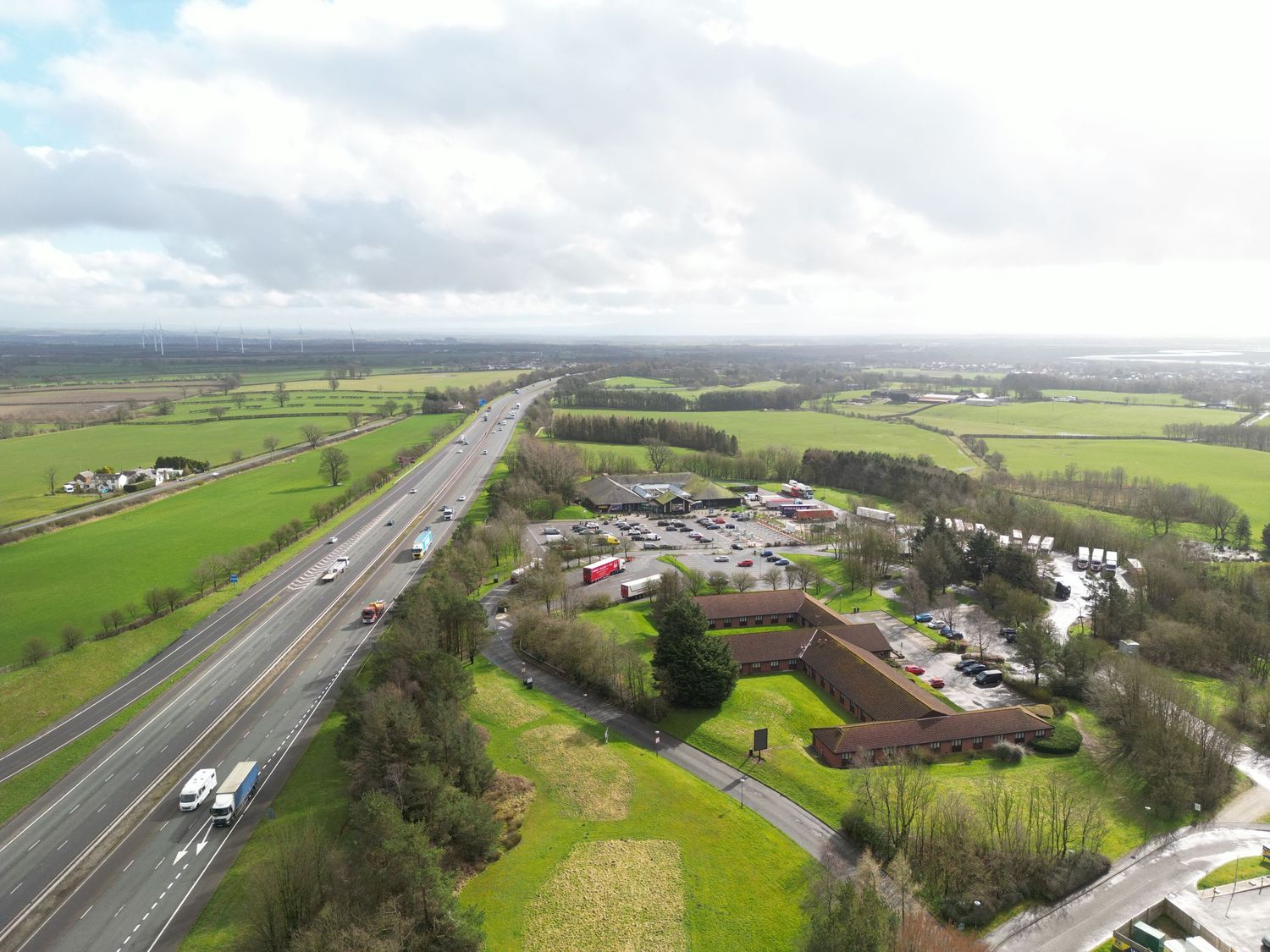 Aerial view of a motorway alongside green fields and a roadside service area under a cloudy sky.