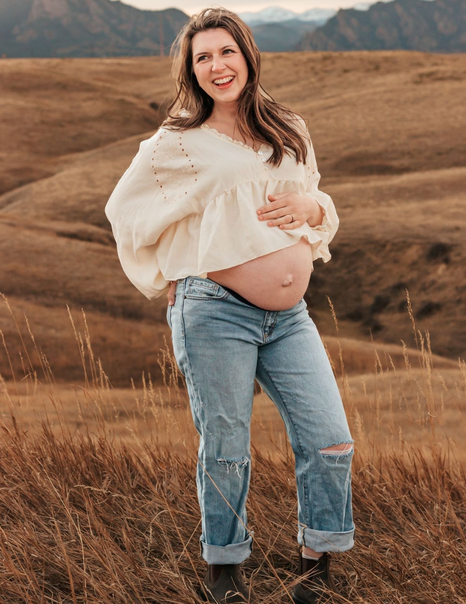 Smiling pregnant woman in cream top and ripped jeans in a golden field with mountain backdrop | Boulder maternity photographer