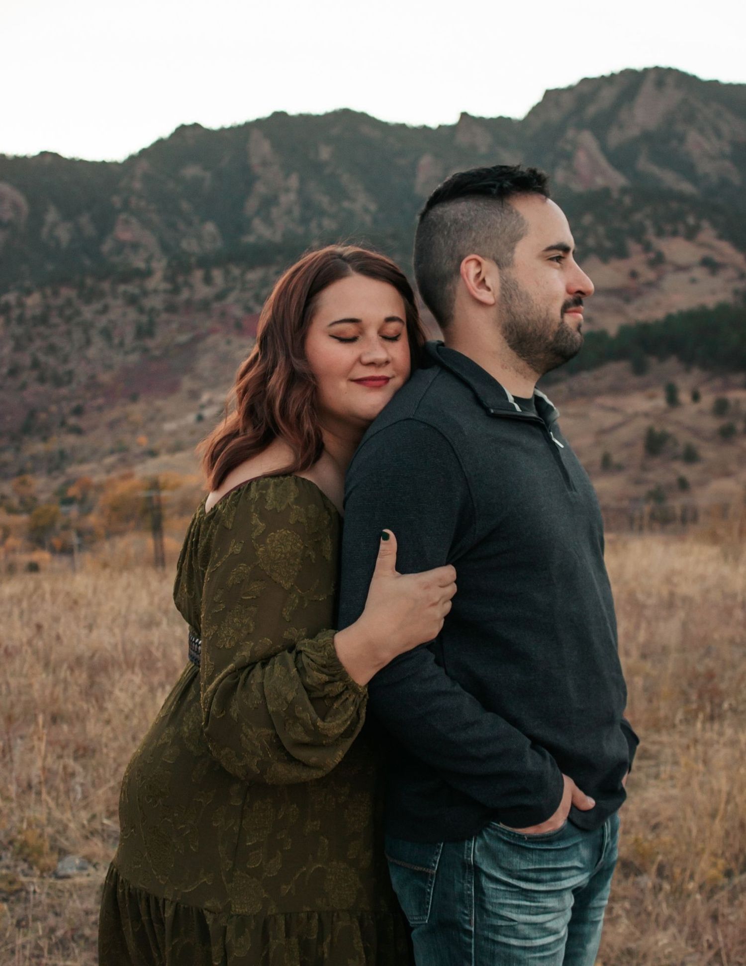 Couple embracing back to back in a field with Rocky Mountain backdrop woman in green dress leaning on man's shoulder | couples photographer
