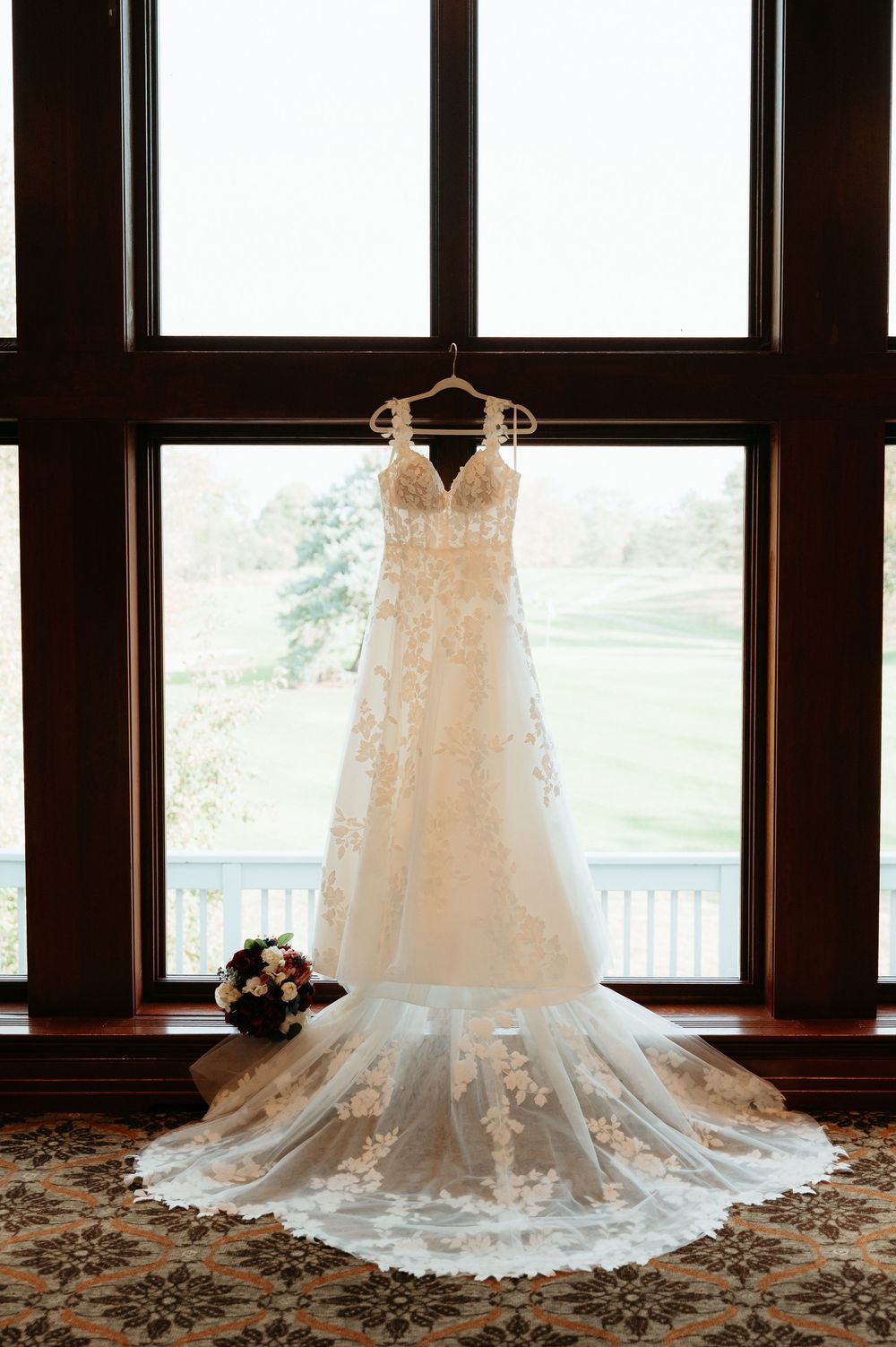 Wedding dress with intricate lace details and long train displayed against bright windows. The wedding dress. Glen Oaks Golf Course, MI.