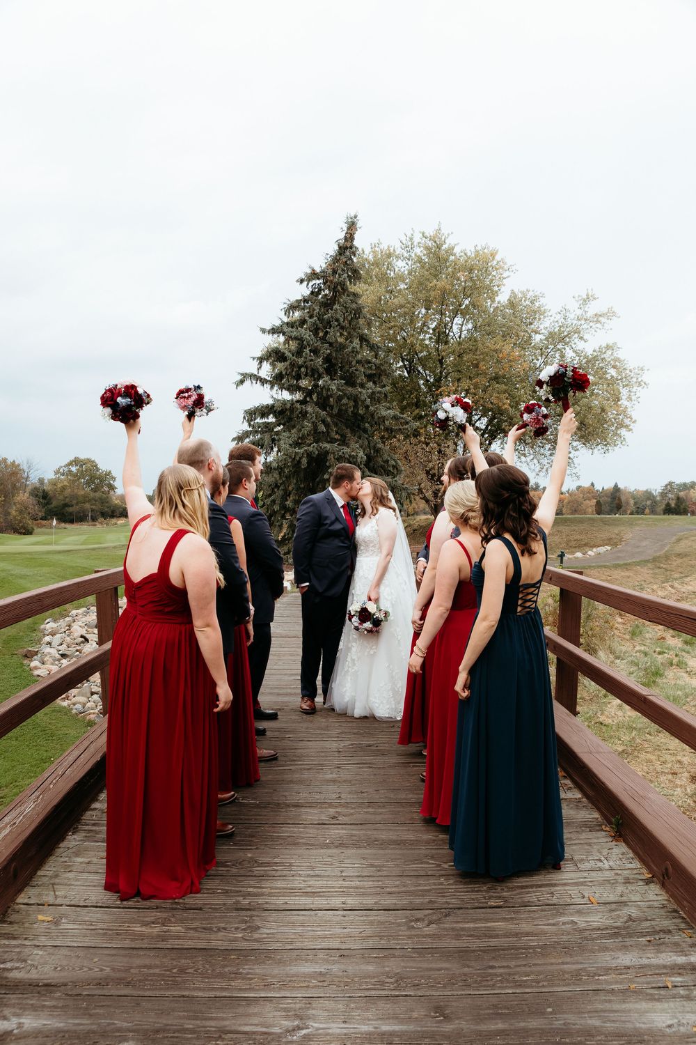 Wedding party in red and navy dresses poses on a wooden bridge. Detroit golf course wedding