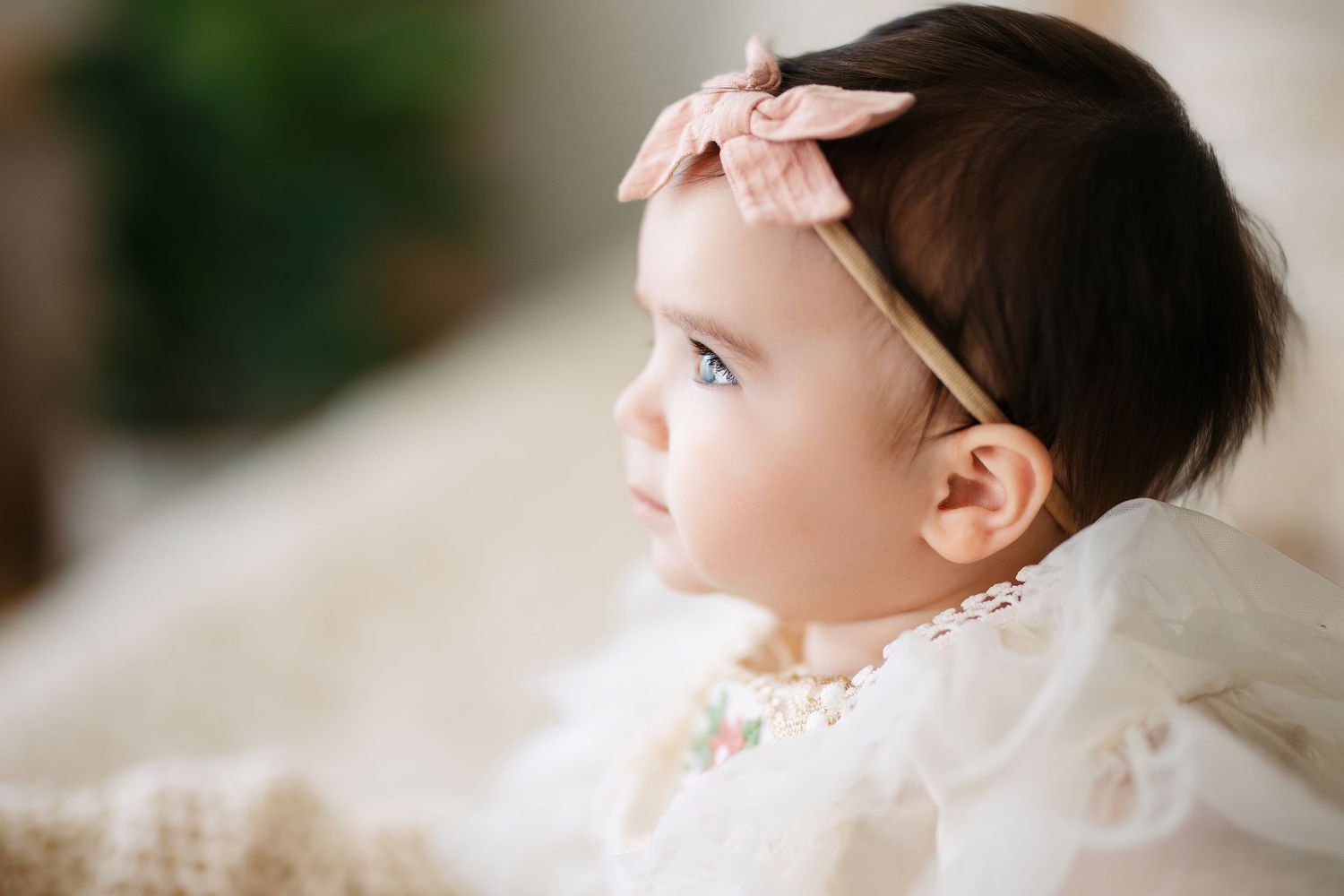 Baby in white lace dress wearing pink bow headband gazes to the side in soft natural light.