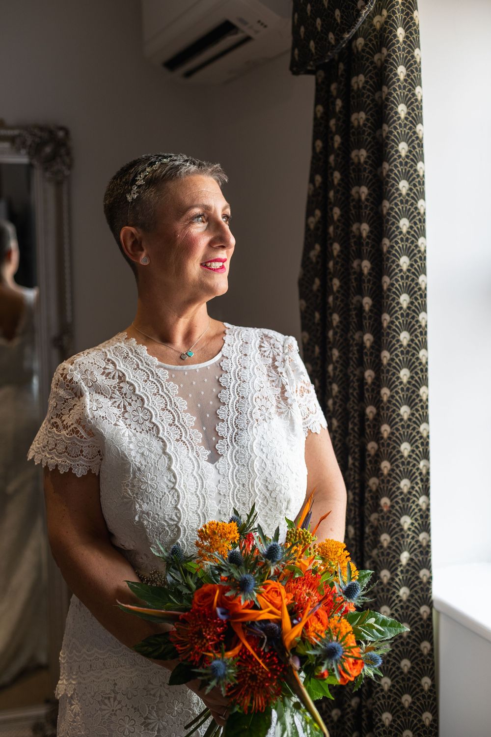 A bride in a white lace dress holds a vibrant orange and red autumn bouquet while standing by a patterned curtain.
