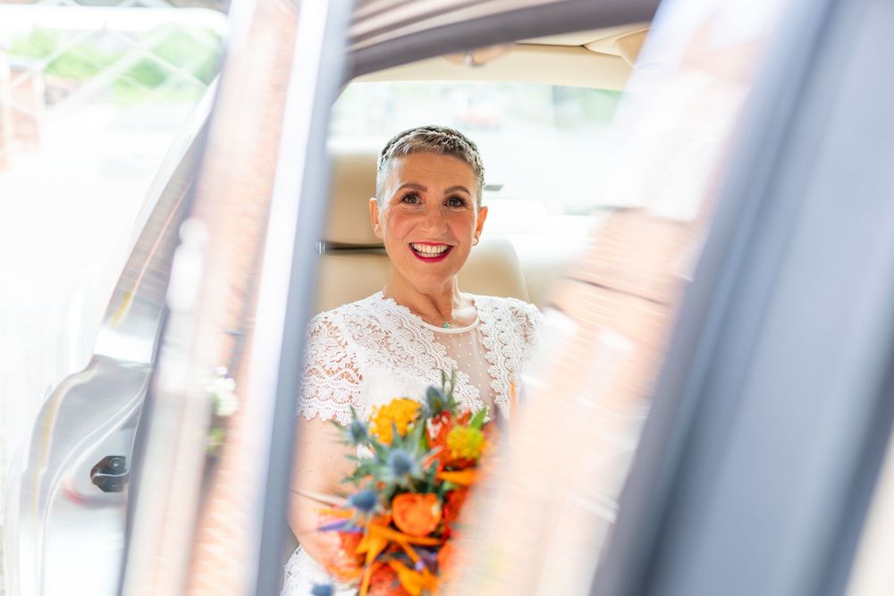 A bride in a white dress and veil holds a vibrant orange and blue bouquet while smiling near a window.