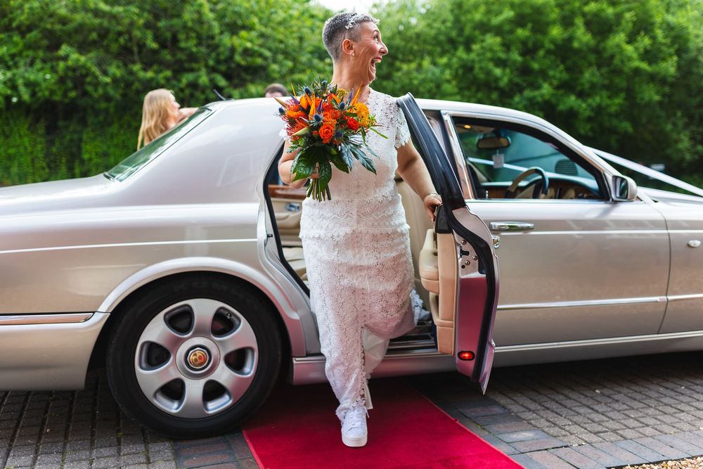 A person in a white dress enters a silver car while holding a bouquet on a brick walkway.