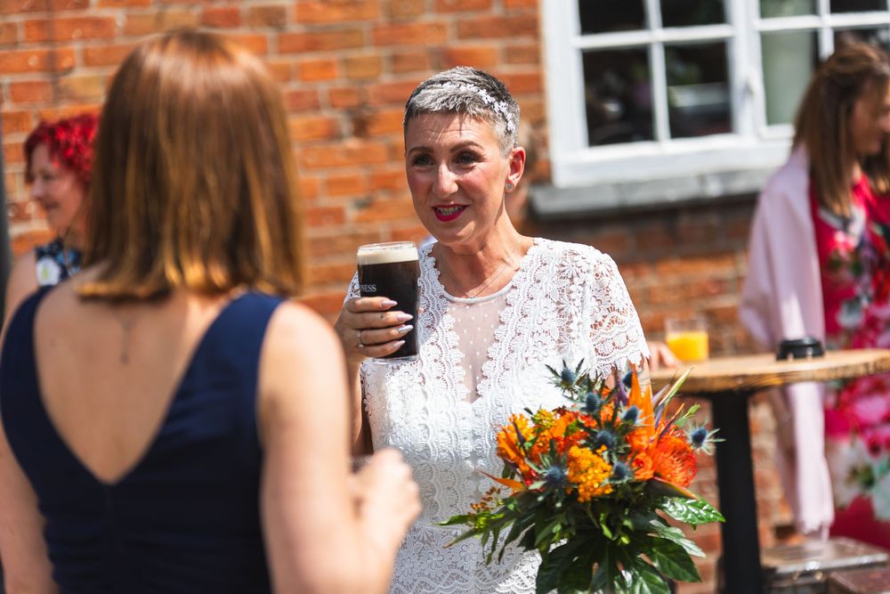 A group of people enjoy a sunny outdoor gathering with colorful flowers and drinks against a rustic brick wall backdrop.