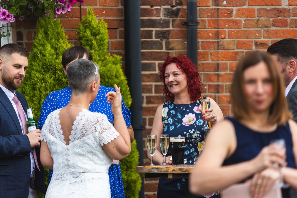 Wedding guests mingling at outdoor reception by brick wall with flowering vines.