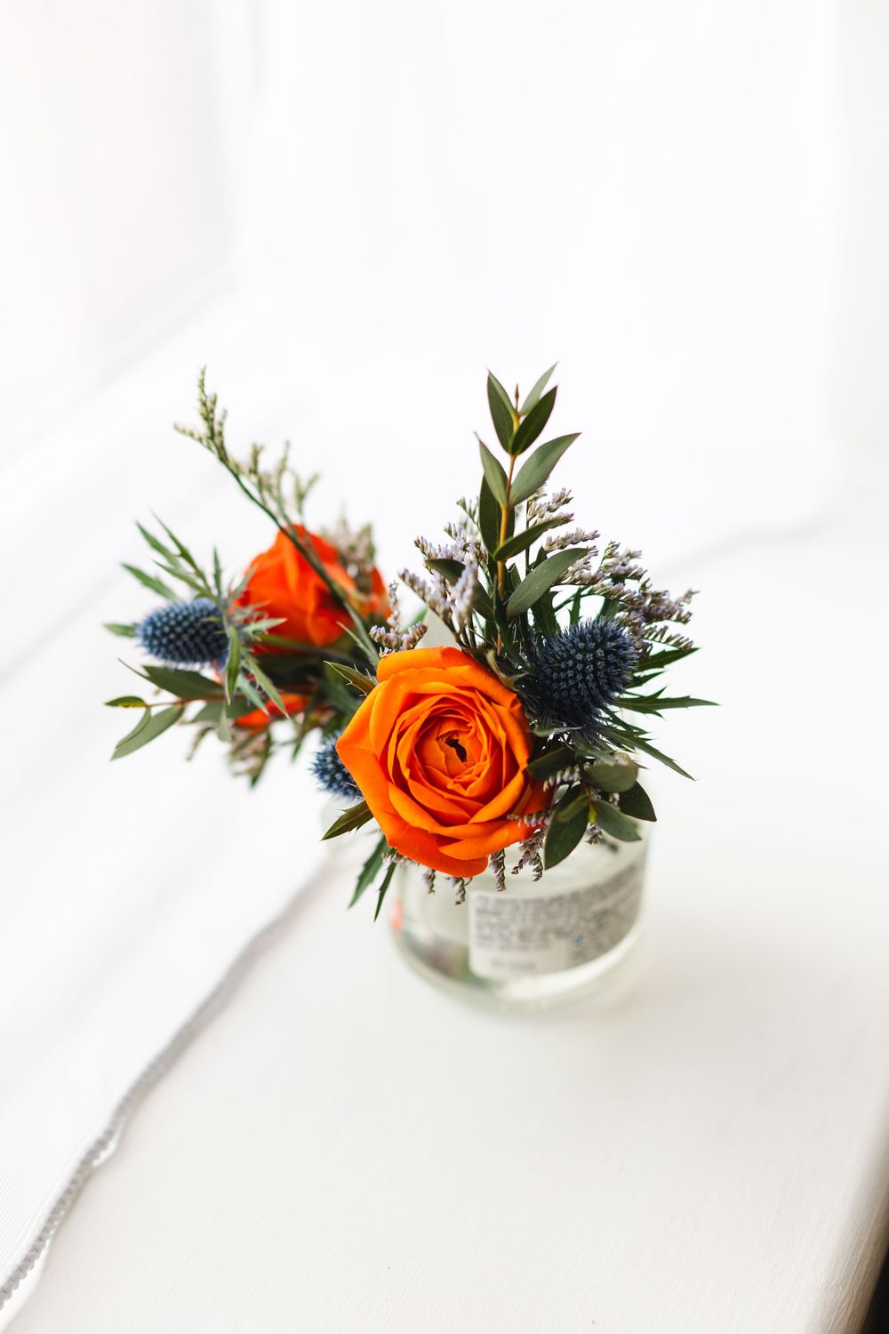 Three delicate bud vases with orange roses and blue thistle flowers arranged on a white surface.