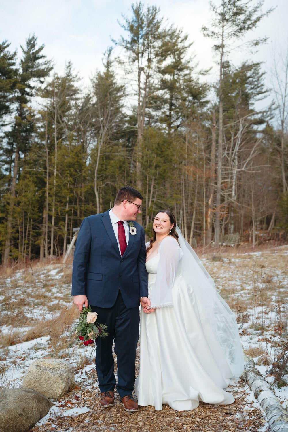 A winter wedding couple shares intimate moments in a snowy forest setting with evergreen trees in the background.