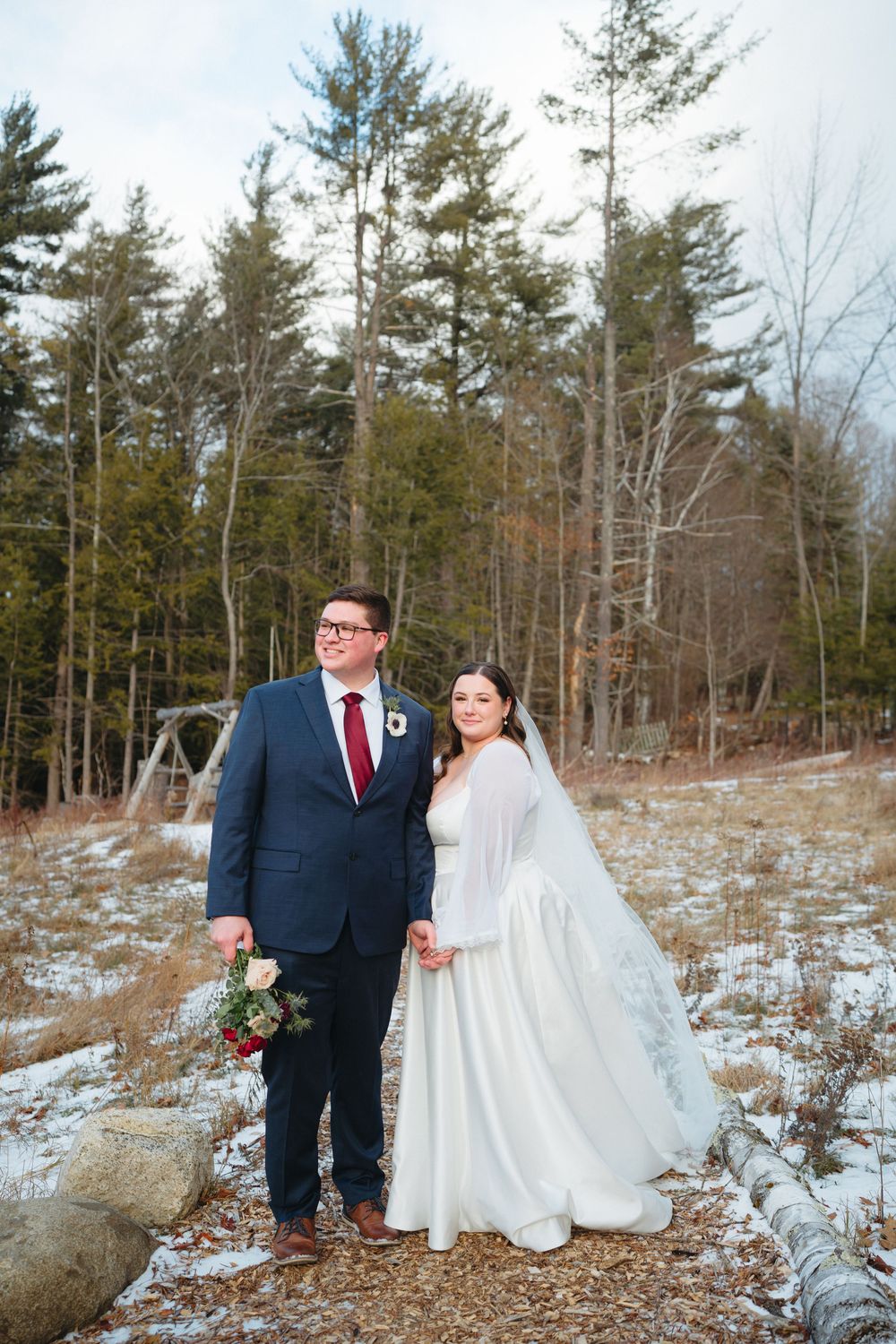 Winter wedding couple poses together in a snowy forest setting with evergreen trees and scattered rocks in the background.