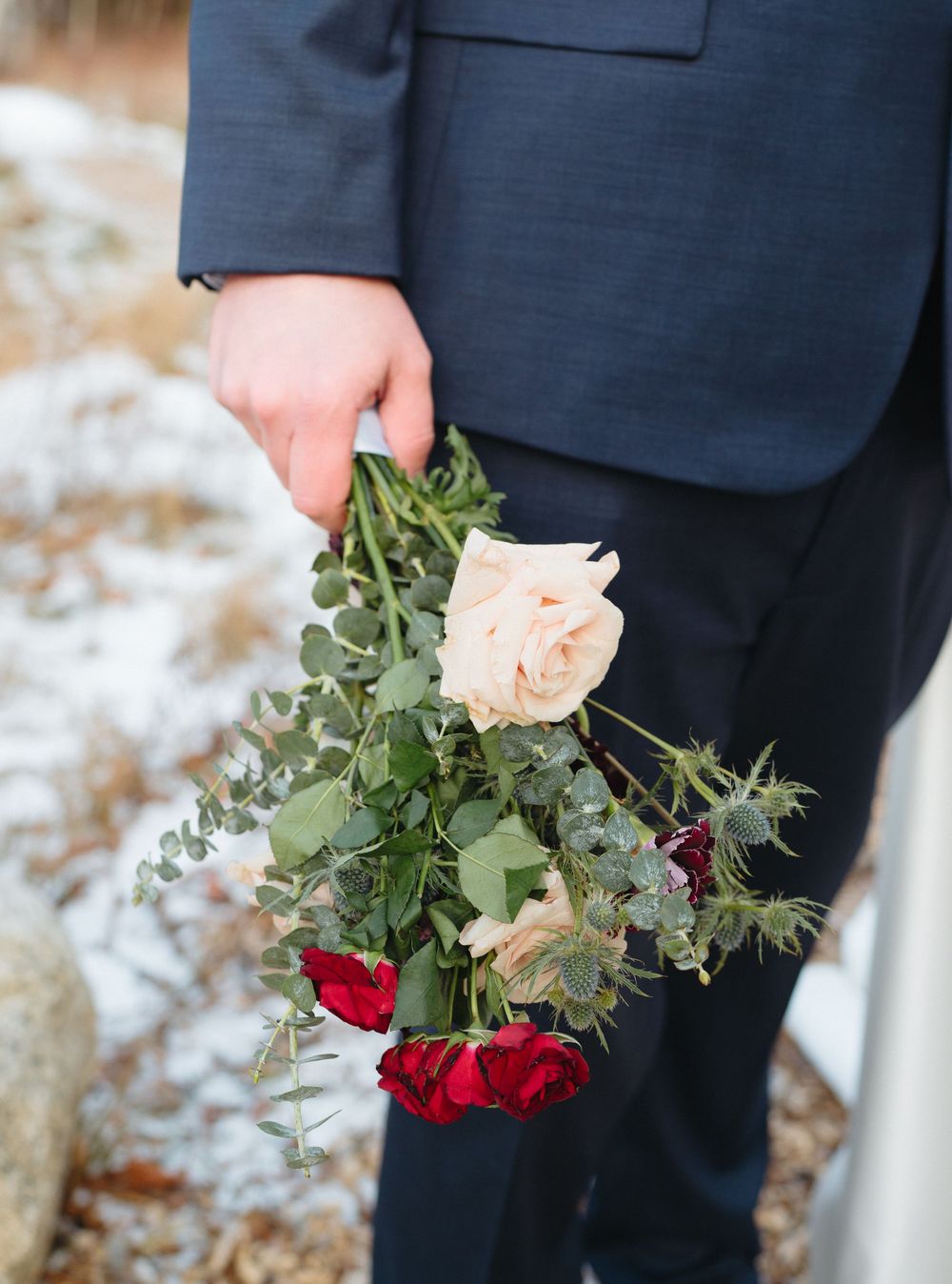 A close-up of hands holding a mixed bouquet with peach roses, red roses and eucalyptus against a snowy background.