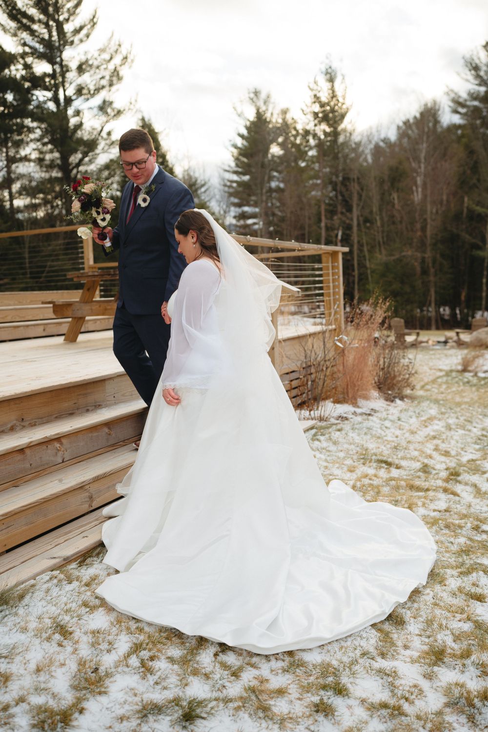 A couple in winter wedding attire share a romantic moment on wooden deck stairs surrounded by snowy evergreens.