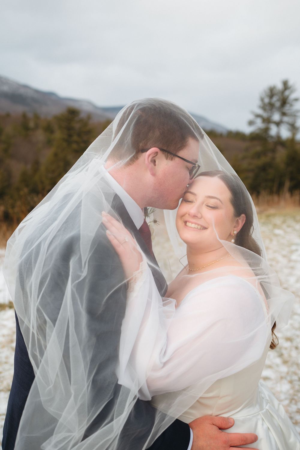 Romantic wedding couple shares intimate moments under flowing veil against scenic mountain backdrop.